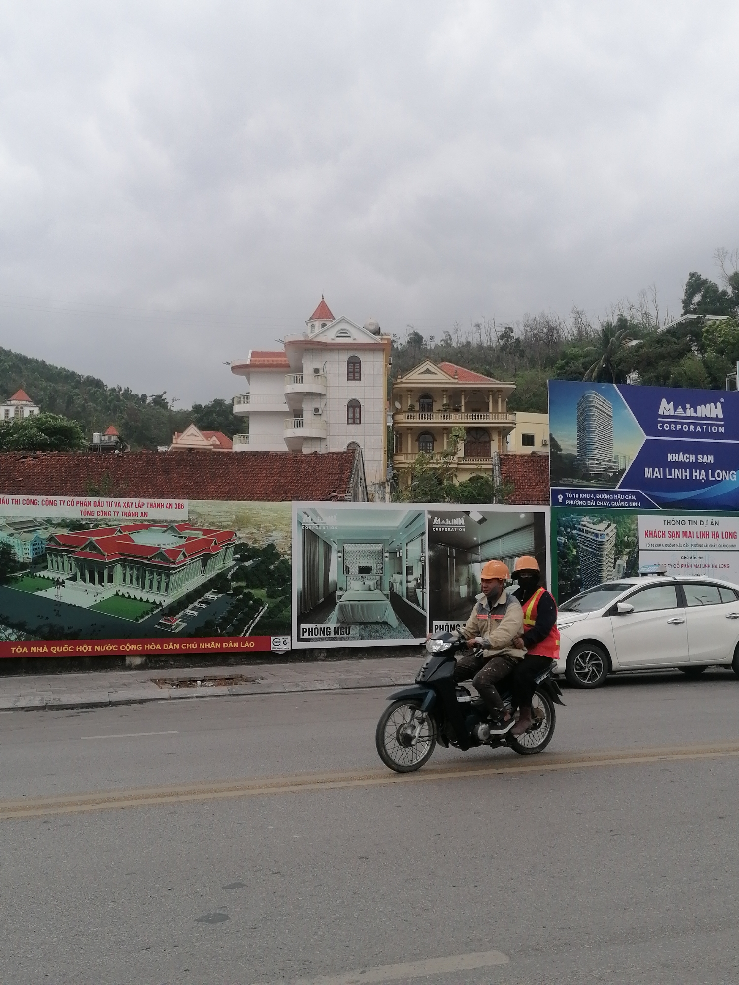 This street view in Bai Chay Ward, Vietnam, captures a moment under an overcast sky, highlighting urban development and daily life. In the foreground, two men, wearing helmets and safety gear including hard hats and an orange vest, are riding a black scooter on an asphalt road. A white Toyota Vios car is partially visible parked or moving alongside.
The midground is dominated by large billboards advertising local construction and development projects. One extensive billboard prominently features a rendering of "TÒA NHÀ QUỐC HỘI NƯỚC CỘNG HÒA DÂN CHỦ NHÂN DÂN LÀO" (National Assembly Building of the Lao People's Democratic Republic), crediting "CÔNG TY CỔ PHẦN ĐẦU TƯ VÀ XÂY LẮP THÀNH AN 386" (Thanh An 386 Investment and Construction Joint Stock Company) for its construction. Adjacent to it, another billboard displays an interior room with the label "PHÒNG NGỦ" (Bedroom). Further right, two more billboards promote "KHÁCH SẠN MAI LINH HẠ LONG" (Mai Linh Ha Long Hotel) by "MAILINH CORPORATION," providing an address in "PHƯỜNG BÃI CHÁY, QUẢNG NINH."
Behind the billboards, several multi-story residential buildings with distinctive red-tiled roofs and balconies are visible, nestled against a backdrop of green, tree-covered hills. The sky is uniformly gray, indicating a dull or cloudy day. The scene depicts a bustling area undergoing construction and development, characteristic of a growing urban landscape.