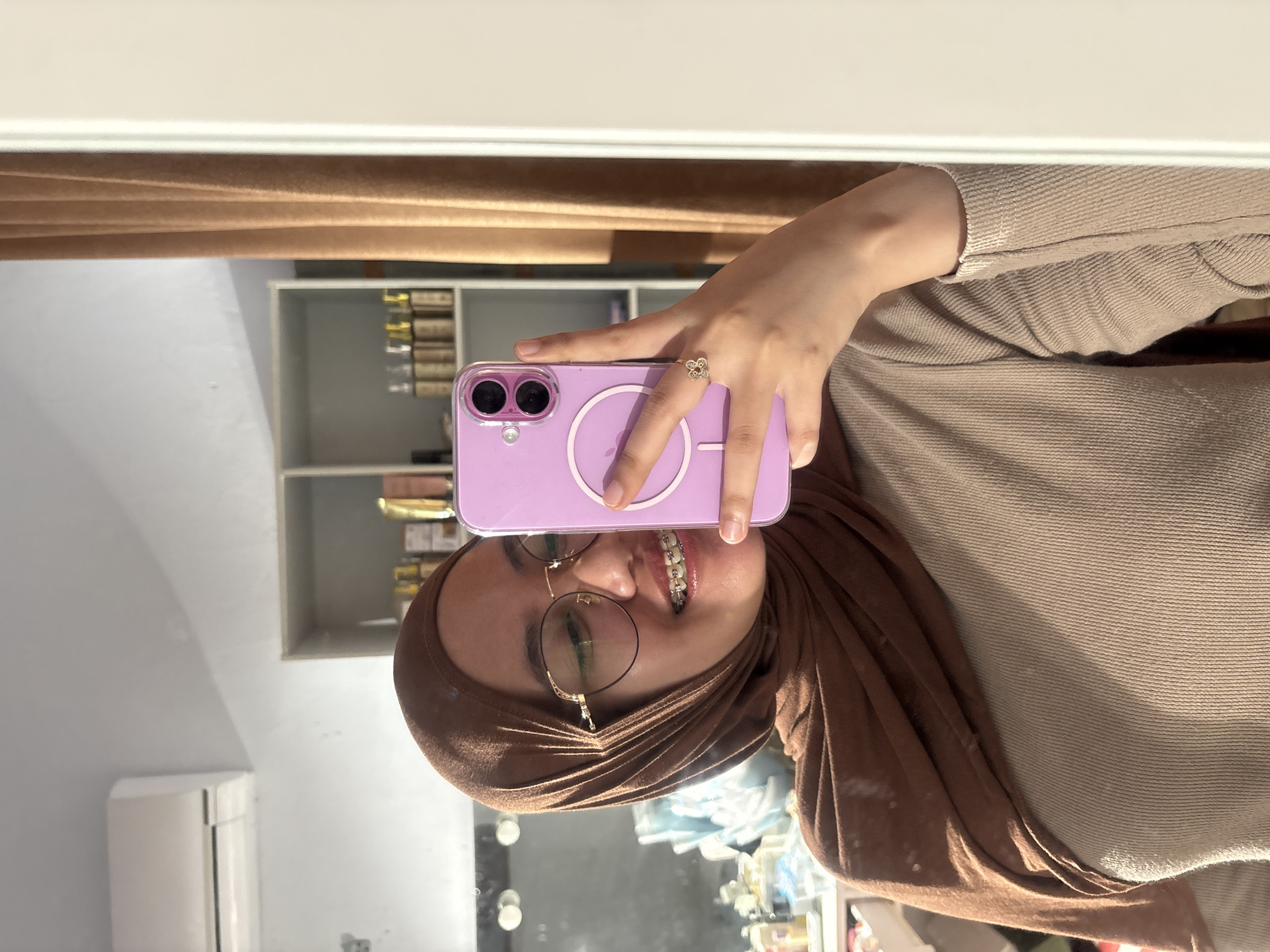 A person with brown hijab and eyeglasses is taking a selfie with a purple phone. The person has braces on their teeth and a flower-shaped ring on their finger. The background shows a shelf with items on it, a beige curtain, and a white air conditioning unit. The image appears to be taken indoors, possibly in a home. There is no verifiable evidence that this image was taken in Gadingrejo, Indonesia.
