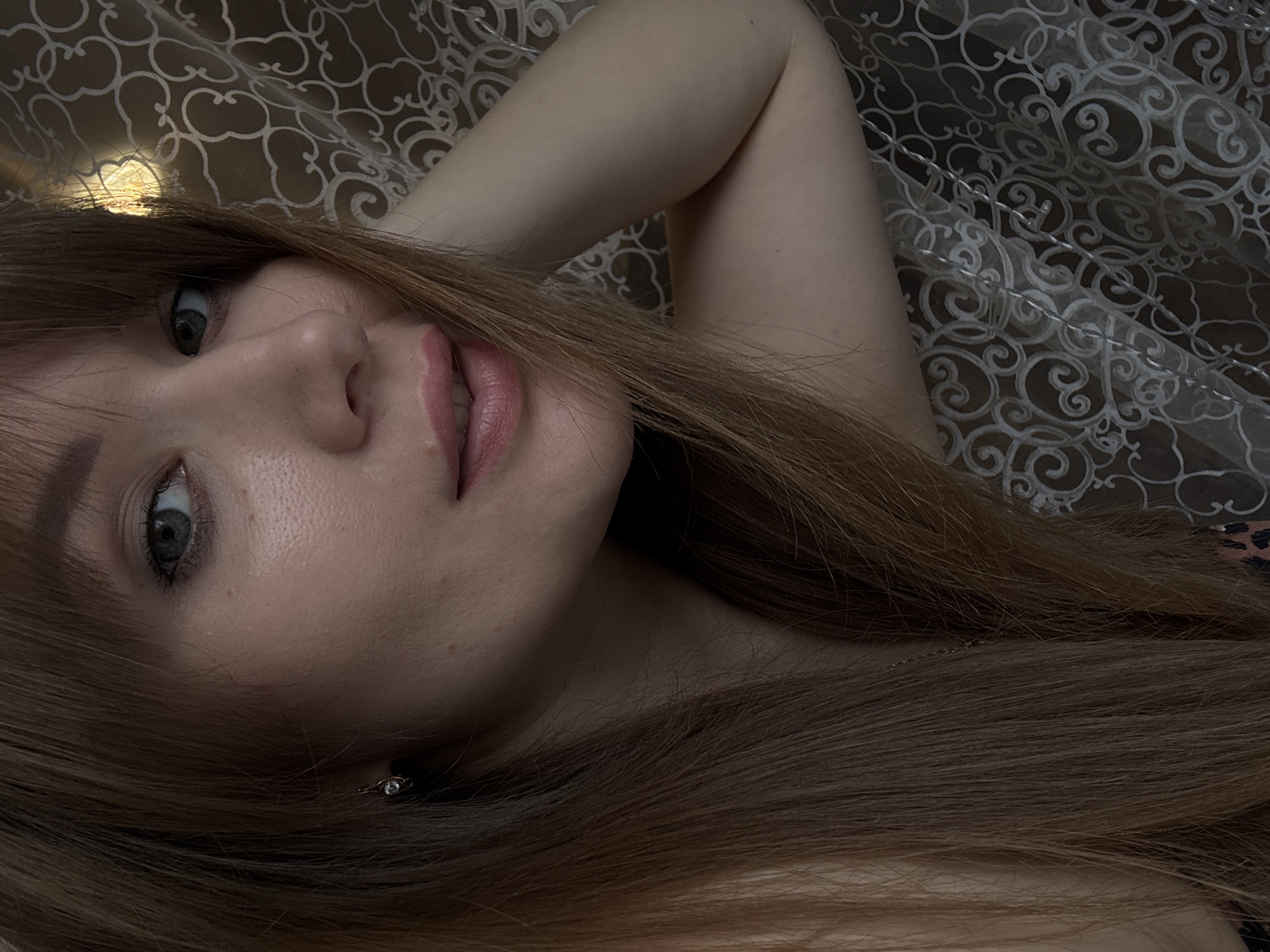 A young woman with auburn hair and green eyes is seen in a close-up shot, lying down with her arm behind her head. She is looking towards the camera with a slight smile. The background consists of a patterned fabric, possibly a curtain or bedding, with a warm light source visible in the upper right corner. The image appears to be taken indoors. There is no information within the image to suggest it is located in Chaikovsky, Russia.