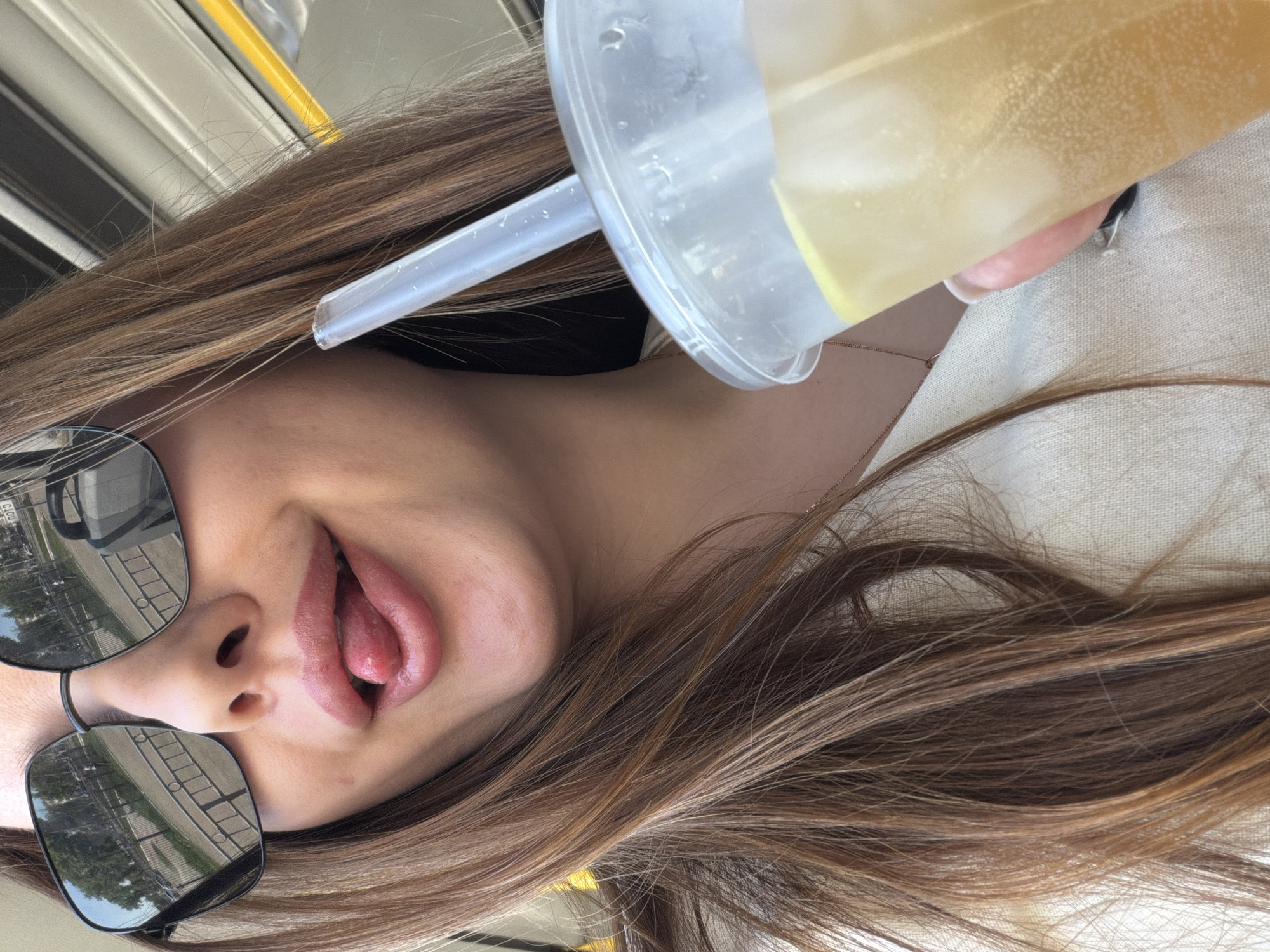 A young woman is posing with a drink, likely in a public transportation setting, possibly the interior of a bus or tram. She is positioned sideways to the camera, holding a clear plastic cup with a straw, suggesting a beverage containing ice and a light-colored liquid, potentially lemonade. Her sunglasses reflect an urban landscape, possibly the city of Chaikovsky, Russia, with buildings, trees, and what appear to be bus shelters or similar structures. The environment is bright, suggesting it is daytime. She is sticking out her tongue. She wears a light-colored top, and a fine chain is visible around her neck. The background includes a yellow handrail, further supporting the public transport location.