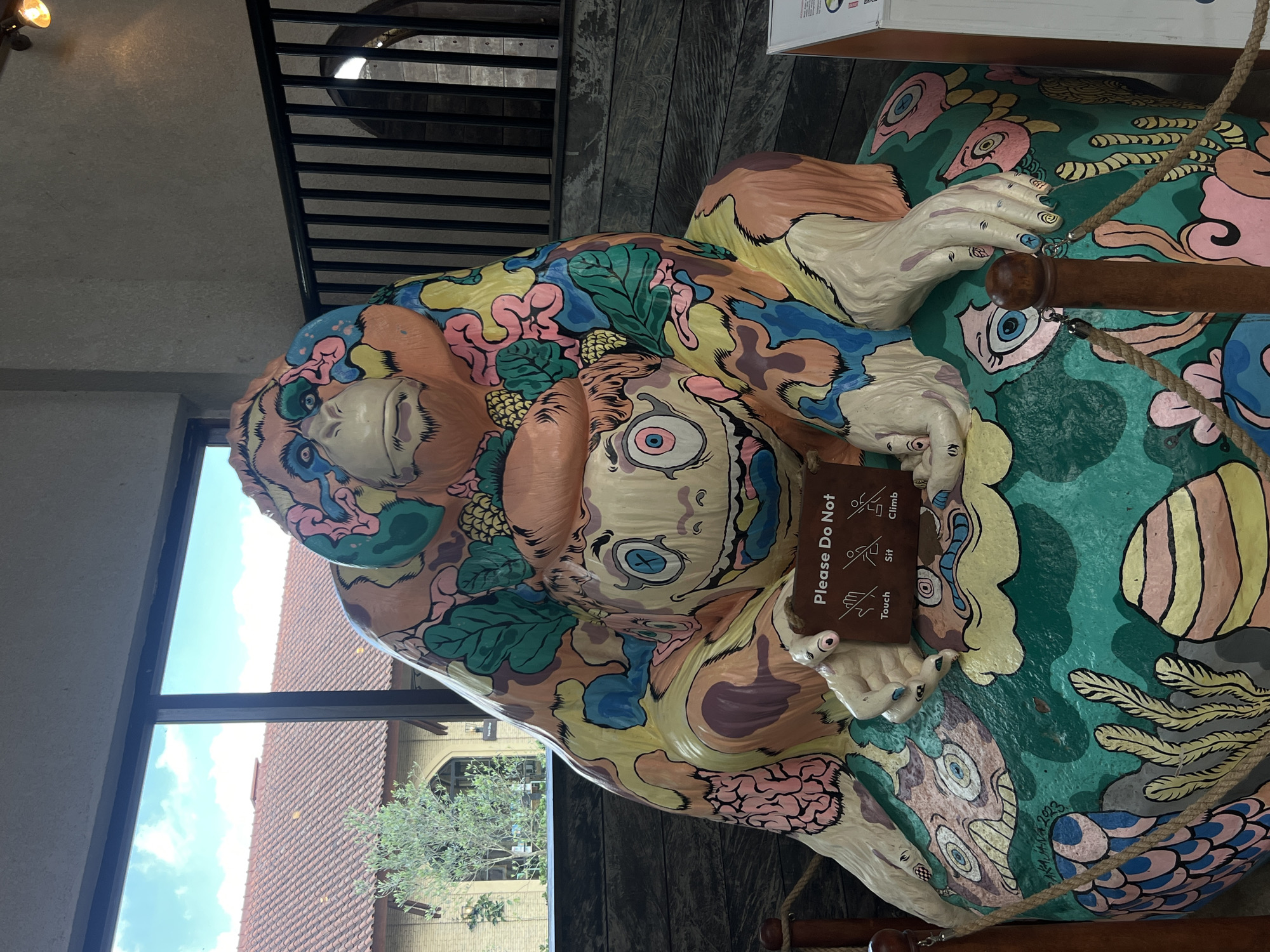 This image shows a colorful statue of an orangutan and a smaller creature sitting on a rocky, underwater-themed base. The statue is located indoors, with a large window in the background showing a building with a red tiled roof and trees outside. A railing is visible behind the statue, and a sign in front of it reads "Please Do Not Touch, Sit, Climb." The orangutan is depicted with vibrant colors and patterns, and the smaller creature has large, wide eyes and a grinning expression. The base of the statue is adorned with painted images of fish and coral. The overall impression is of a whimsical and artistic display. The location is in the city of Sukasada, Indonesia.