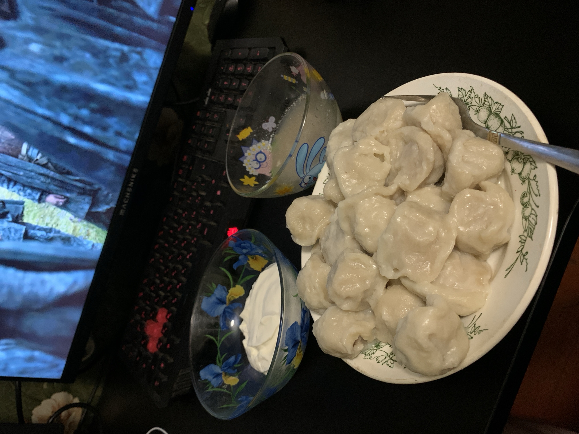 A plate of dumplings, a bowl of sour cream, and a bowl of broth are arranged on a black surface, likely a desk, in front of a computer monitor displaying a scene from a medieval-themed program. A keyboard with red-backlit keys is visible in the background. The dumplings are light-colored and appear to be of the varenyky or pelmeni variety. The sour cream is thick and white, filling one of the decorative glass bowls. The broth in the second glass bowl is translucent with some lighter colored solids. The plate of dumplings features a green floral pattern around its rim. The overall scene suggests a casual meal enjoyed while watching content on a computer, with no specific indicators of a city or location.