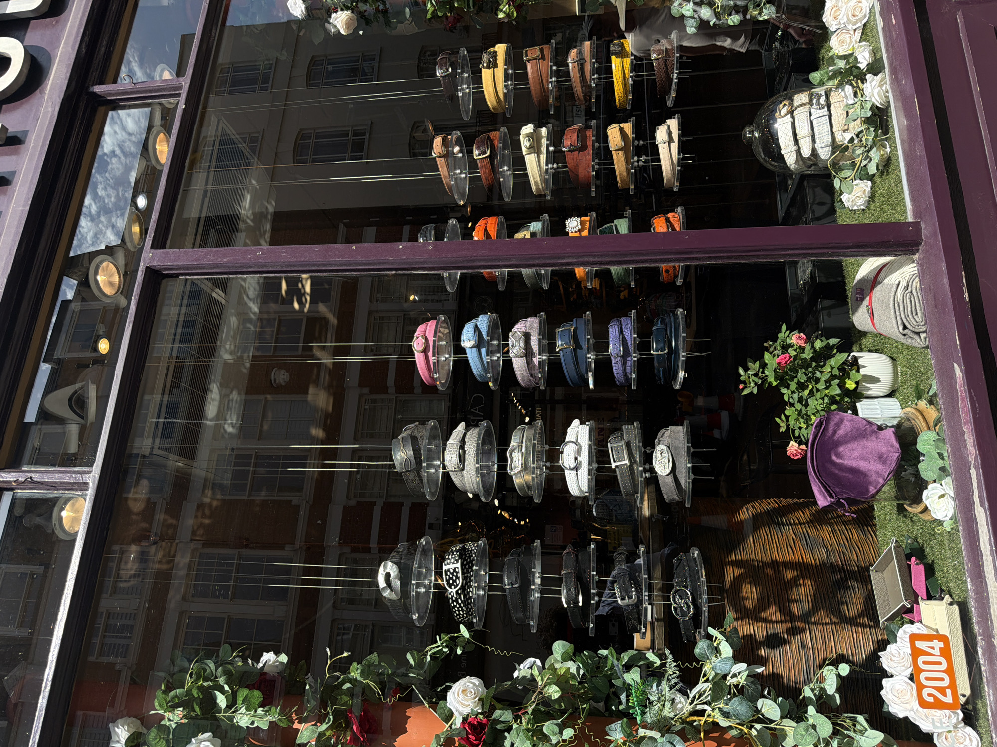 A display of watches is viewed through a shop window in London, United Kingdom. The watches are arranged in rows, each watch supported by a clear plastic stand, and they vary in color and style, ranging from pink to brown. Floral arrangements, including white and red roses, are placed around the watches, adding to the aesthetic of the window display. A reflective surface behind the watches shows the facade of the buildings across the street, revealing the urban environment. A purple bonnet is placed near a container. A sign with "2004" in orange and white is near the bottom right of the display.