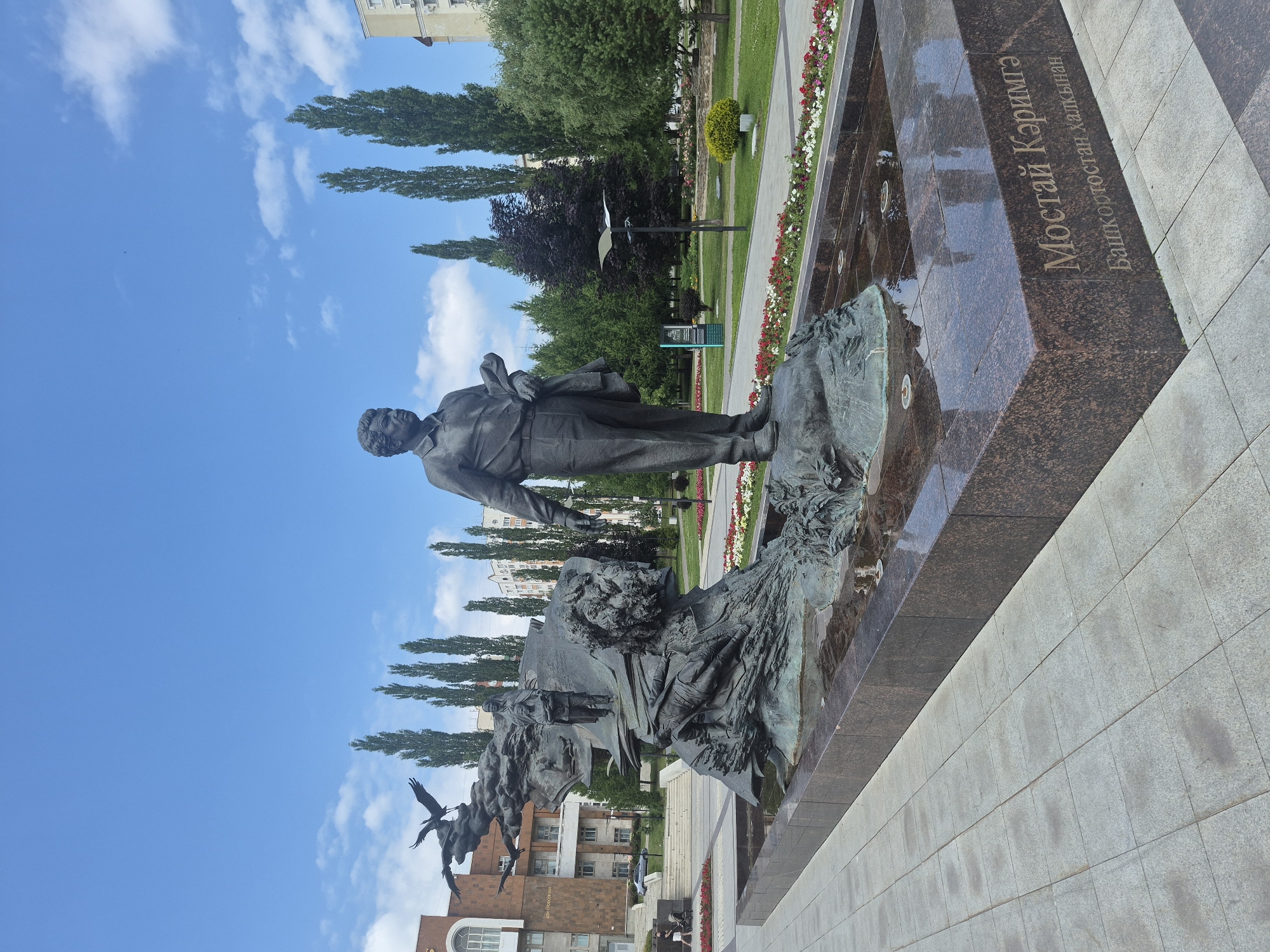 This is a full-shot, eye-level, outdoor photograph of a large bronze monument in Ufa, Russia. The monument, located in a park setting, features a central statue of a man in a suit, holding a jacket over his arm and a book in his other hand. He stands on a rocky base. To the left of the man is a more complex sculptural element depicting figures and birds in flight, emerging from what appears to be abstract forms. A fountain or water feature surrounds the base of the monument. The monument is set on a paved walkway with flowerbeds lining the edges. In the background, there are tall, slender trees, a building with a facade of red brick and large windows, and other residential buildings. The sky is blue with scattered white clouds. Text is visible on the granite base of the monument, identifying it as "Мостай Кэримгә" and "Башкортостан халкынан".