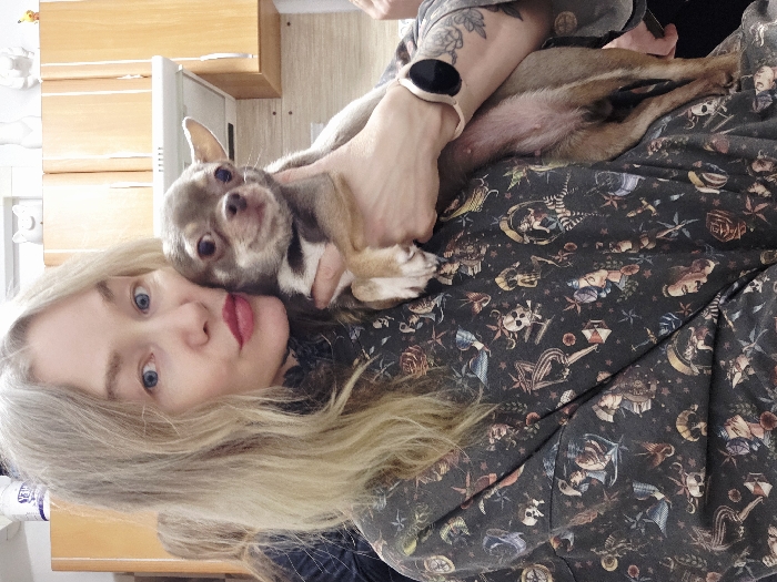 A woman with blonde hair and blue eyes poses with a small chihuahua. The dog rests on the woman's chest, held by her hand. The woman wears a dark patterned shirt, and a smartwatch is visible on her wrist. The background includes a kitchen cabinet and a light-colored wall. The image is rotated counter-clockwise.