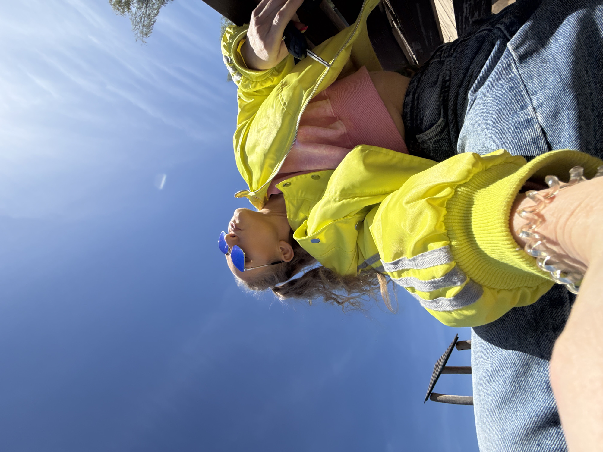 A low-angle shot depicts a person wearing blue jeans, a pink sweatshirt, and a bright yellow jacket with reflective stripes on the sleeves. The jacket is partially zipped. The person is also wearing sunglasses with blue lenses. The background is a clear blue sky with some faint clouds. The person appears to be sitting on a wooden structure, possibly a bench or railing, as suggested by the wooden slats visible in the upper and lower portions of the frame. The location is not discernible from the image.