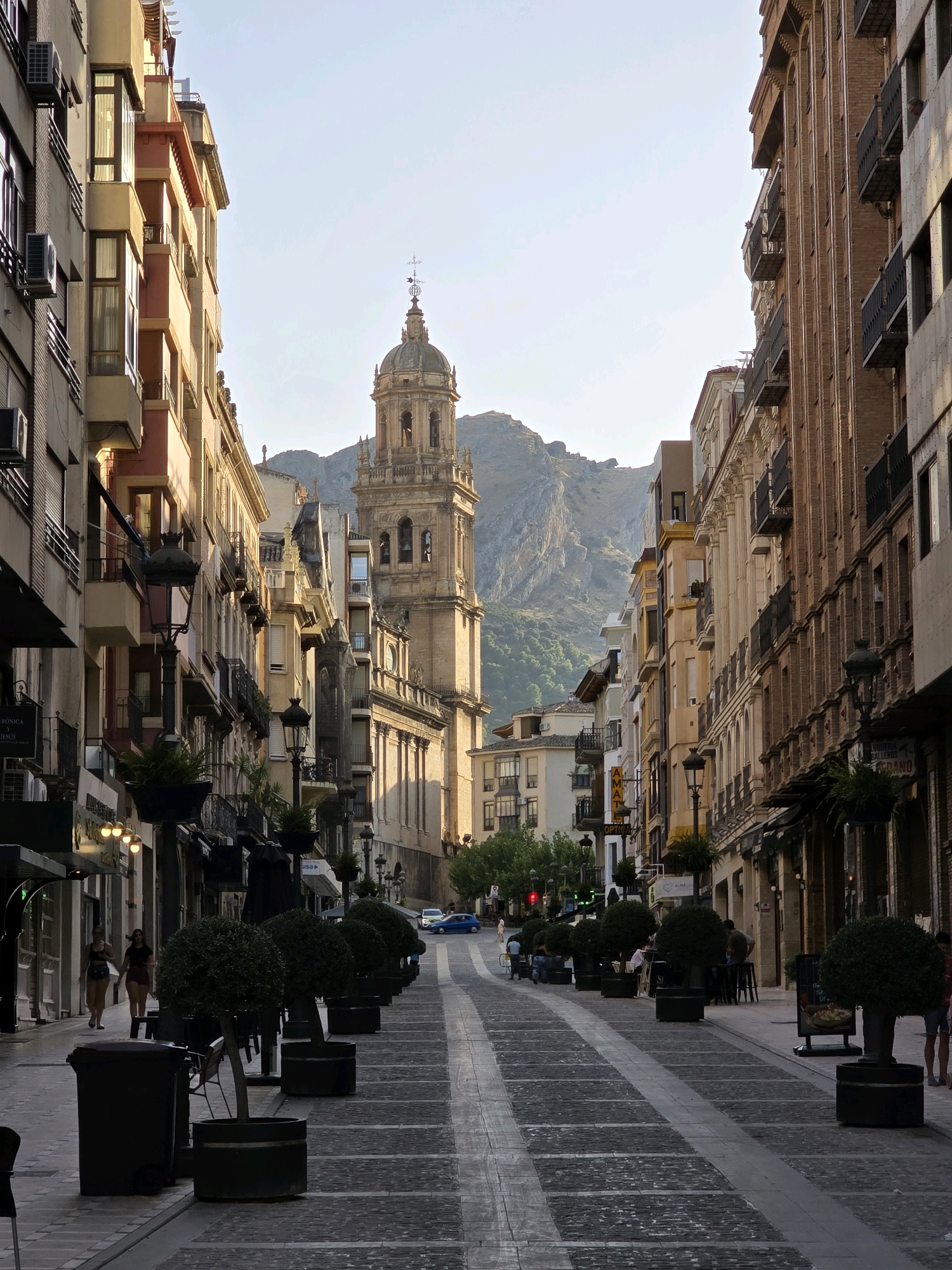 This is a street-level photograph taken in Jaén, Spain, looking down a pedestrianized avenue flanked by multi-story buildings. A prominent church tower with arched openings and a dome-like structure is visible in the distance, framed by the buildings and a rugged mountain landscape. The street is paved with cobblestones and a central lighter-colored strip. Planters with small, rounded trees are lined up along the avenue, and lampposts are present. Several people are walking on the sidewalks and some are seated at outdoor tables. A blue car is visible further down the street. The buildings exhibit architectural details such as balconies, windows, and awnings, with some displaying commercial signage. The sky is clear and bright, indicating daylight.