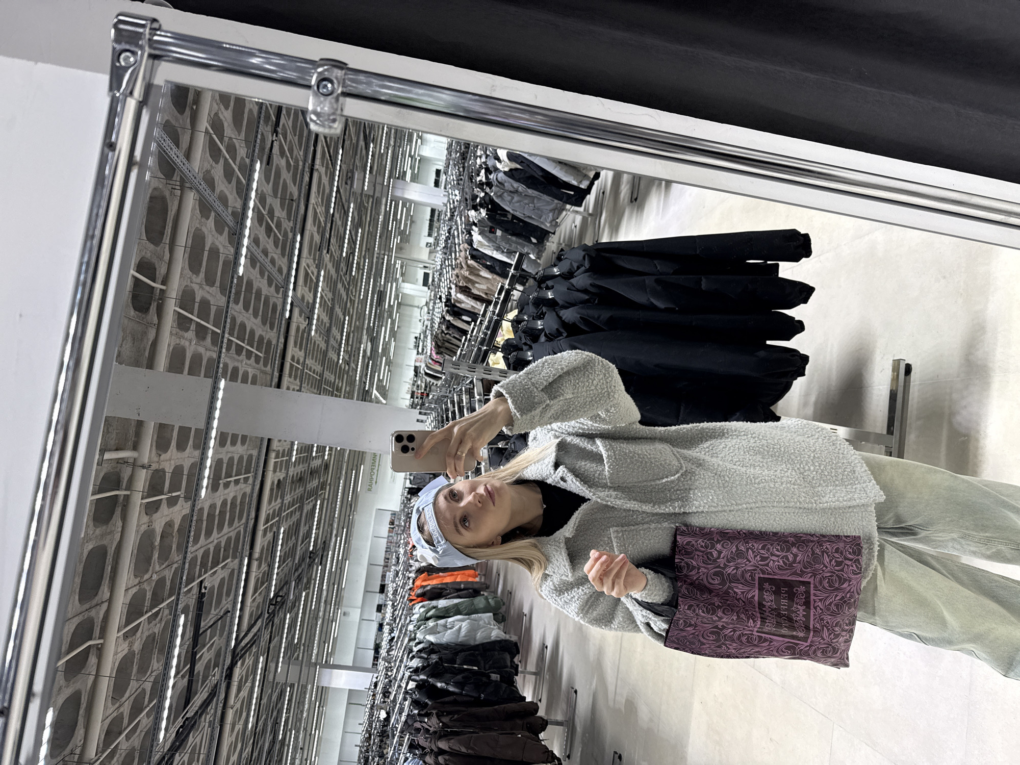 The image captures a young woman taking a selfie in a mirror within a large retail environment, possibly a warehouse store, due to the extensive rows of clothing racks and high ceilings. The woman is holding a smartphone and is dressed in a light-colored jacket and pants. She is wearing a light-colored head covering and carrying a purple shopping bag with a decorative pattern. The store's interior features rows of clothing on racks, organized by color and type. The high ceilings are industrial with exposed ductwork and lighting fixtures. Based on the bag's logo, the store is likely located in Izhevsk, Russia.