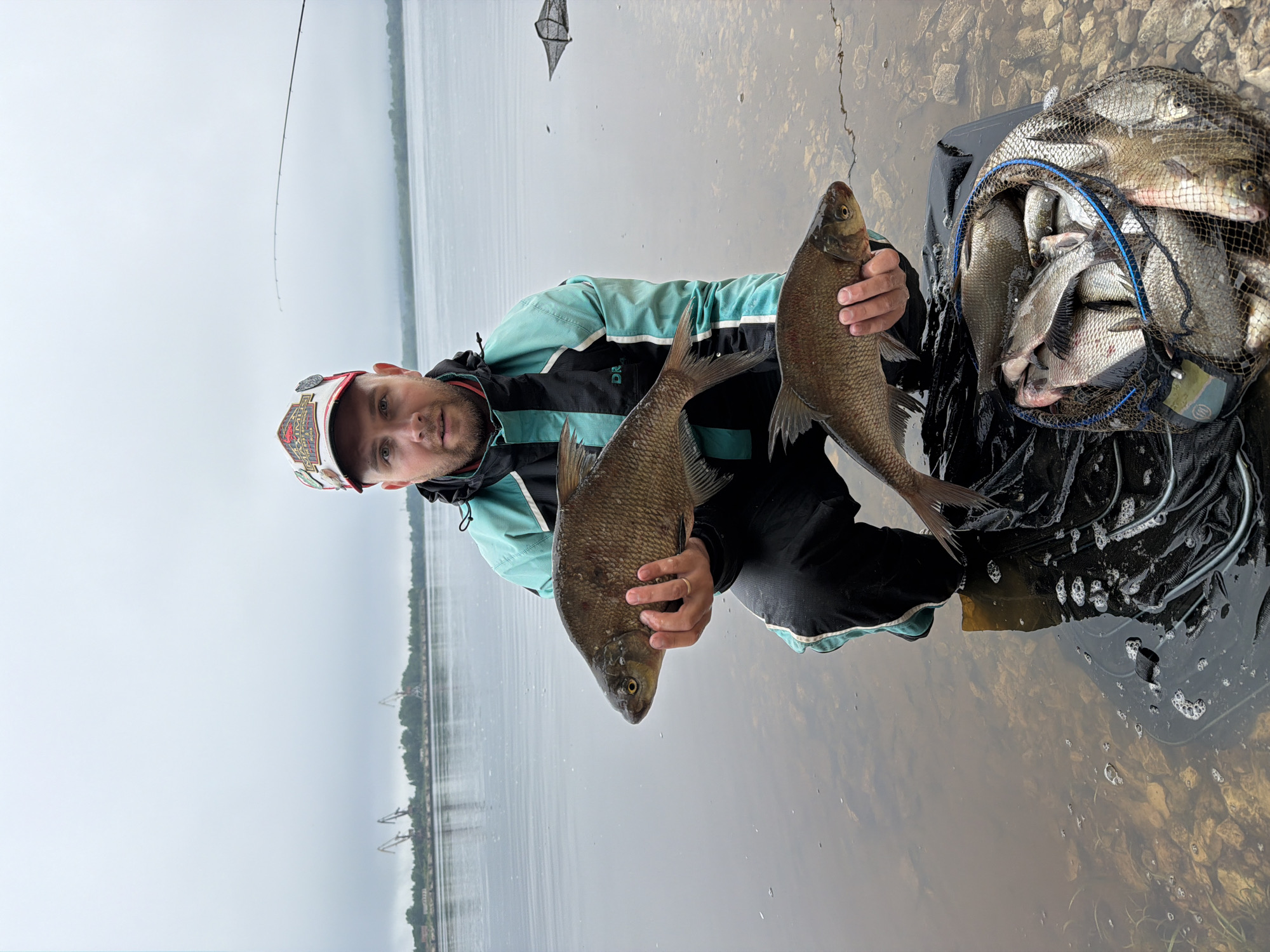 The image, taken in Chaikovsky, Russia, captures a man proudly displaying his fishing catch. He stands in shallow, clear water with a rocky bottom, holding two large, silver-scaled fish, likely bream, in his hands. The man has a beard and is wearing a baseball cap with various patches, a turquoise and black jacket (with "DR" visible on it), and black pants. His expression is neutral, looking directly at the camera.
To the man's right, a black fishing net or basket is partially submerged in the water, overflowing with numerous smaller fish of the same species, indicating a successful fishing trip. A fishing rod extends upwards and into the distance on the left side of the frame.
The setting is a large body of water, possibly a river or reservoir, under an overcast, grey sky. The water is calm and reflects the sky. A distant shoreline with trees and some indistinct industrial structures, possibly cranes or buildings, is visible across the water. The overall lighting suggests a cloudy day. The scene depicts the satisfying outcome of a day spent fishing.