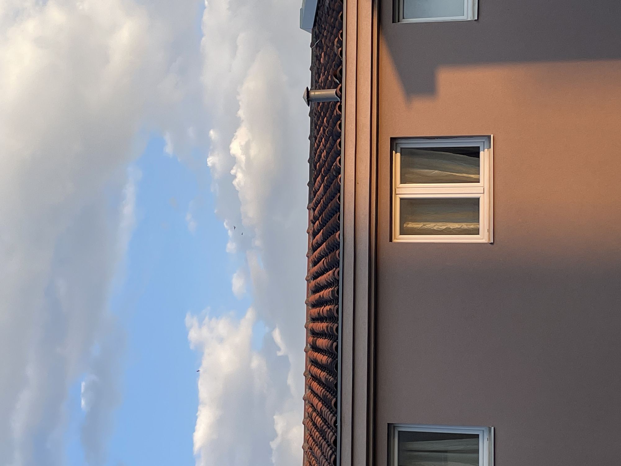 A low-angle, eye-level shot captures the side of a building with a tiled roof, under a sky of blue and white clouds. The building is painted in a light brown hue, with two windows visible, one on top of the other. The windows appear to have white frames and curtains or blinds inside. The roof is covered in red-brown tiles, with a ventilation pipe extending from it. A shadow stretches across the building's wall. The location of the image is in Bar, Montenegro.