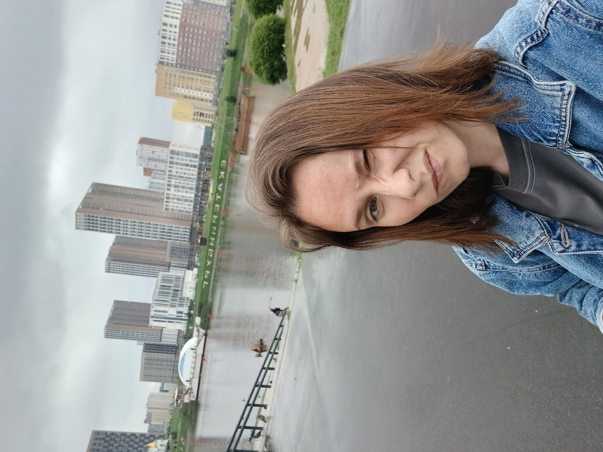 In Yekaterinburg, Russia, a woman with brown hair, wearing a denim jacket over a grey t-shirt, is taking a selfie. She is winking and has a playful expression on her face. The background shows a waterfront with a body of water, a ramp leading down to it with railings, and several ducks on the ramp. Across the water, a cityscape of modern high-rise buildings is visible under a cloudy sky. A sign with the Cyrillic word "ЕКАТЕРИНБУРГ" (Yekaterinburg) is visible along the waterfront. There are also some trees and green spaces in the distance.