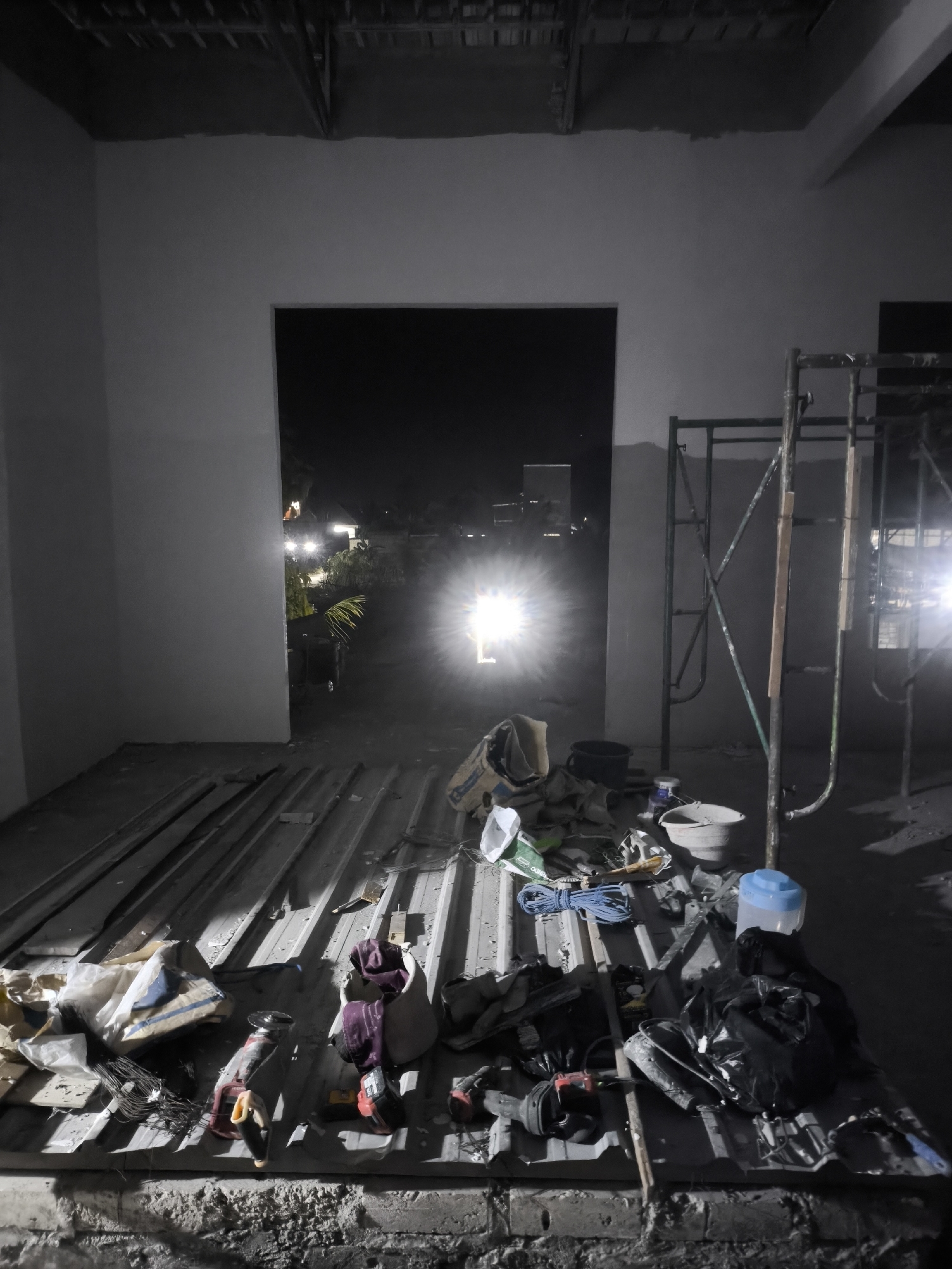 An indoor construction site at night is depicted. A doorway opens to an outdoor area illuminated by bright lights, possibly from vehicles or equipment, creating lens flare. The indoor space is in disarray with construction materials and tools scattered across a metal-ribbed floor. Visible items include scaffolding, buckets, bags of materials, coiled rope, power tools like angle grinders and drills, and various other debris. The walls are unpainted, and the ceiling structure is exposed. The overall impression is one of an active, yet unfinished, construction project at night.