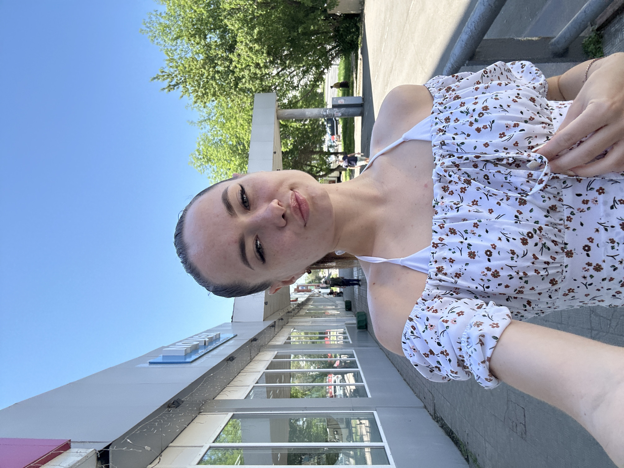 A young adult female is depicted in a selfie, taken outdoors on a sunny day in the city of Unknown, Unknown. She has light skin, dark hair pulled back, visible freckles across her nose and cheeks, and prominent eyelashes. Her lips are slightly pursed, and she wears a white off-the-shoulder top adorned with a small brown and pink floral pattern, revealing a white halter-neck strap underneath. Her left hand is seen adjusting a tie-front detail on her top, and a delicate chain bracelet is on her left wrist.
She stands on a gray paved sidewalk. To her left is a long, low-rise commercial building with a gray facade and multiple large rectangular windows reflecting green foliage. A partial, unreadable sign with blue and white elements is visible above the windows, along with strung decorative lights. Beyond the sidewalk and building, a street or parking area is visible. In the background, numerous green deciduous trees stand under a clear, bright blue sky. Several indistinct figures are present in the background: one person in a dark shirt walks away from the camera on the left, and other blurred individuals are visible further down the street and near the trees on the right. A tall grey pillar structure and a grey metal railing are also present in the background on the right side.