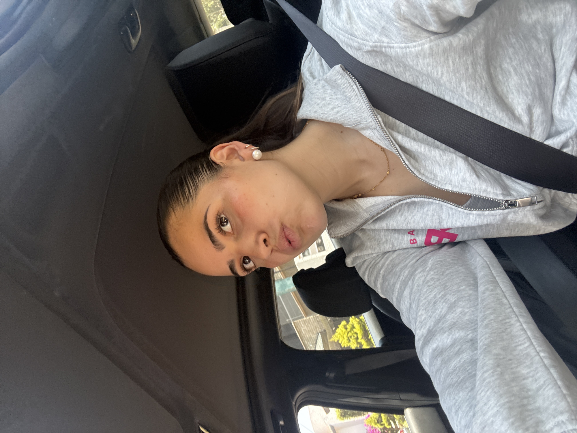 A young woman is captured in a close-up, selfie-style photograph, seated in the passenger side of a car. Her head is tilted to the side, and she looks directly at the camera with a playful, slightly pouty expression. She has dark hair pulled back from her face, light brown eyes, and appears to be wearing subtle makeup, including blush and mascara. She is adorned with a pearl stud earring and a small, delicate ear cuff, along with a thin gold-colored necklace.
The woman is wearing a light grey, zip-up hoodie. Part of a red logo, showing the letters "BA," is visible on her hoodie, suggesting the brand Balenciaga. A black seatbelt is fastened across her chest, and a black car headrest is visible behind her. The car's dark interior ceiling and window frames are also visible. Through the car windows, bright daylight illuminates an outdoor scene featuring green foliage, indicative of trees or bushes, and some building structures, suggesting an urban or suburban setting. The bright light indicates it is daytime. The provided context places the location in San Bernardino Tlaxcalancingo, Mexico.
