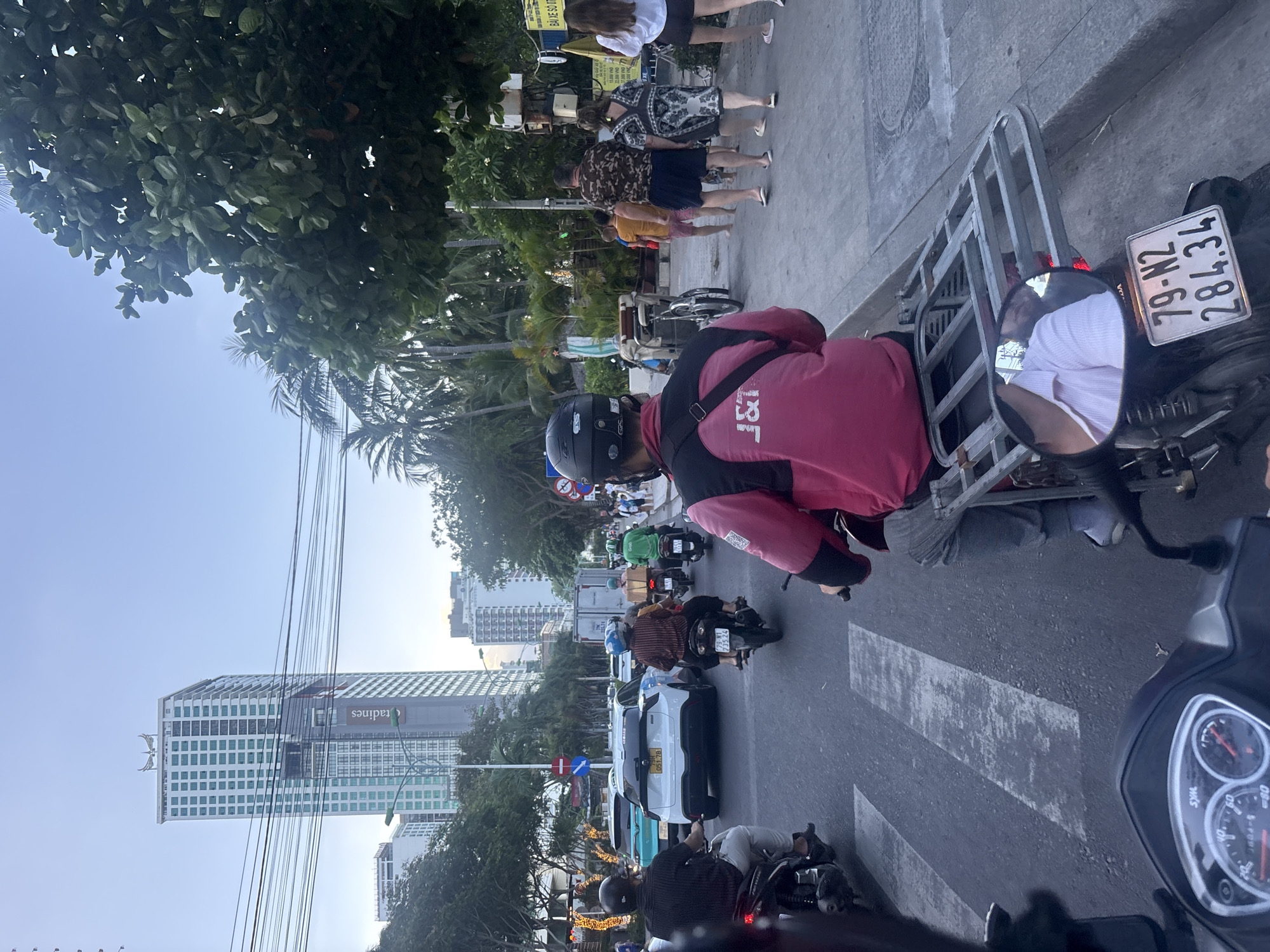 The image depicts a street scene in Nha Trang Ward, Vietnam, during daylight. The perspective is from a scooter in traffic, with several other scooters and a few cars visible on the road. Several pedestrians are on the sidewalk to the right. Tall buildings dominate the distant skyline, including one identifiable structure marked with "Radines". A scooter in the foreground carries a small metal rack, and its license plate reads "79-N2 284.34". The scooter driver directly in front wears a pink J&T Logistics jacket. Trees line the street, partially obscuring the buildings.
