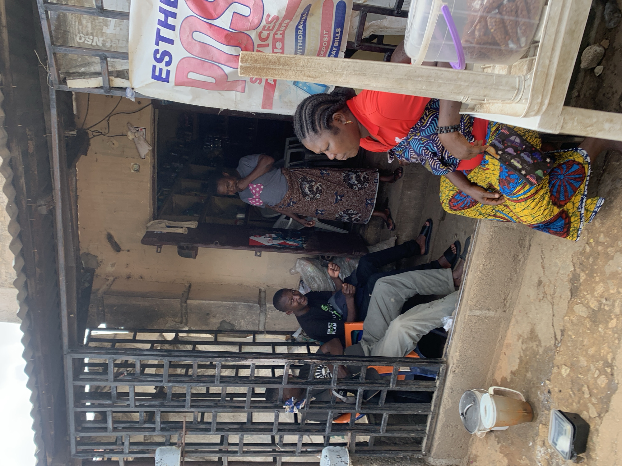 This image captures a vibrant street scene in Jos, Nigeria, under bright daylight. The setting appears to be outside a small commercial establishment or service point, characterized by concrete structures and a visible corrugated iron roof on the left.
In the mid-ground, a woman with braided hair, dressed in a red top and a vibrant patterned skirt (yellow, blue, and red), is bending forward, focused on something out of frame. She holds a small, patterned purse and wears a black beaded bracelet. Above her, a clear plastic container filled with snacks, possibly groundnuts or biscuits, rests on a white surface, alongside a wooden plank.
Standing slightly behind the first woman is another woman wearing a grey t-shirt with pink polka dots and a patterned brown skirt. Further into the background, partially obscured by a dark metal security grille, two men are visible. One man, wearing a black t-shirt with "EQUITY PLAN" printed on it, is seated on an orange plastic stool, looking towards the right with a relaxed expression. The legs of another man, clad in light grey pants, are also visible behind the grille.
A large, partially visible white banner with red and blue lettering hangs prominently, bearing text such as "ESTHE," "ROS," "SERVICES," "WITHDRAWAL," "DEPOSIT," and "PAY BILLS," indicating the nature of the business. Other fragments like "OSN" (reversed) and "T" are also discernible. The ground is a mix of worn concrete and dirt. Several plastic containers are scattered on the ground in the lower right. The overall impression is one of everyday community life and commerce in an urban or semi-urban environment.