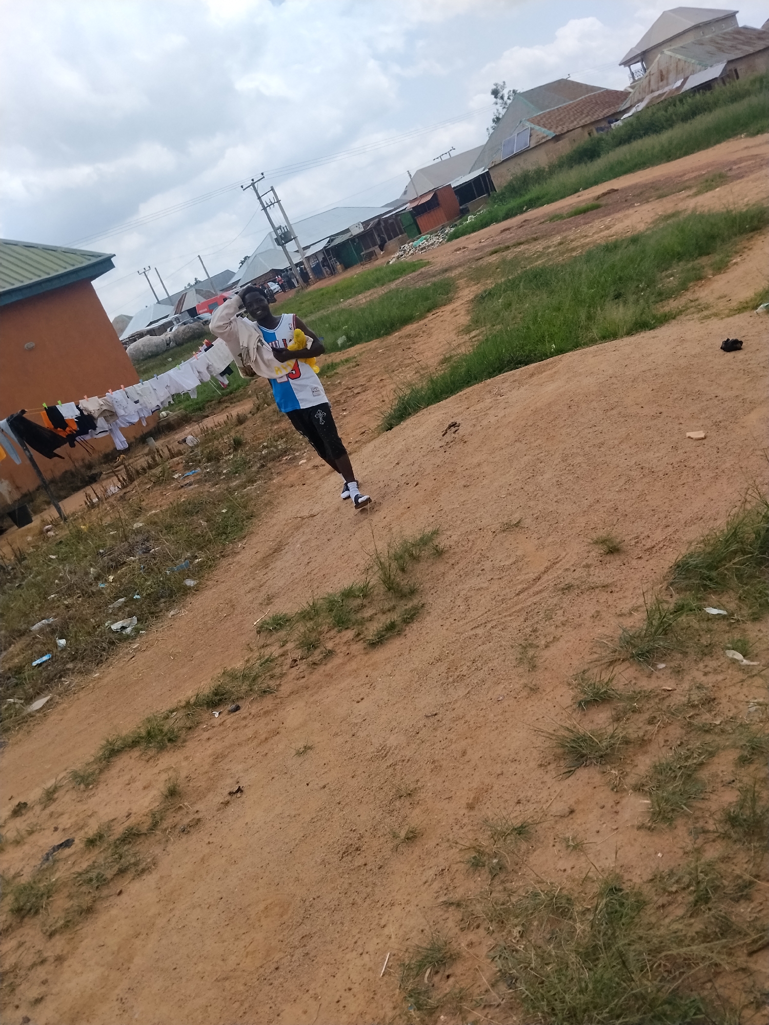 In Durumi, Nigeria, a young Black male stands on a dirt path, holding a yellow stuffed toy and a bundle of white clothing. He is wearing a white, blue, and red basketball jersey with black shorts and white sneakers. Behind him, laundry hangs on a clothesline stretched between two poles, with a brown building to his left. The background features several buildings with corrugated metal roofs and a cloudy sky. The ground is a mix of dirt and sparse green grass, with scattered debris.