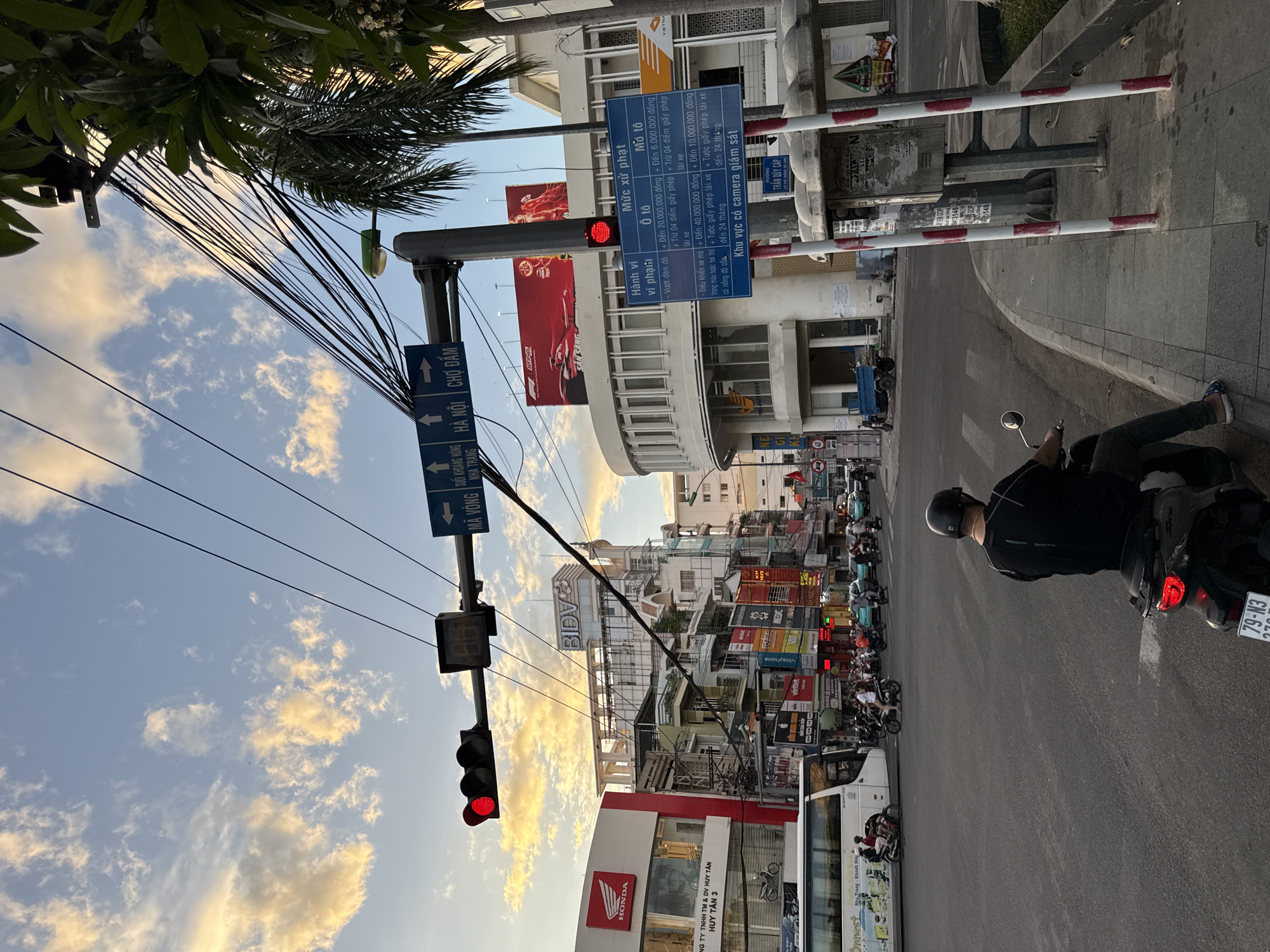 The image captures a street scene in Nha Trang, Vietnam, during what appears to be either dawn or dusk, given the warm lighting and dramatic clouds in the sky. A traffic light signals red. Several signs are visible, including directional signs indicating "MÃ VONG SUỐI KHOÁNG NHA TRANG", "HÀ NỘI", and "CHỢ ĐẦM". Other signs with Vietnamese text are also present, one of which lists traffic violation fines.
On the street, numerous motorbikes and a white bus are moving through the intersection. A person wearing a black helmet is seated on a motorbike in the foreground, facing away from the camera. The buildings lining the street are a mix of modern and older structures, with various shopfronts and advertisements. Power lines are heavily concentrated overhead. The sidewalk features a bus stop shelter and traffic poles. A palm tree is visible on the right side of the frame.
