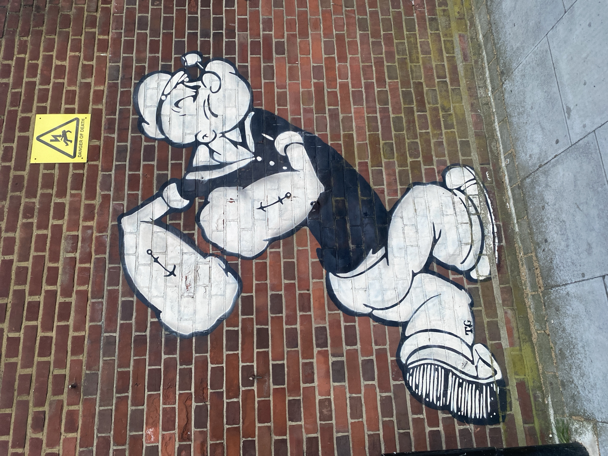 Popeye the Sailor Man is depicted in graffiti on a brick wall next to a Danger of Death sign. The wall adjoins a paved sidewalk. The graffiti shows Popeye with clenched fists, anchor tattoos, and his traditional sailor's outfit. The letters "TG" appear near his shoe. The location is London, United Kingdom.