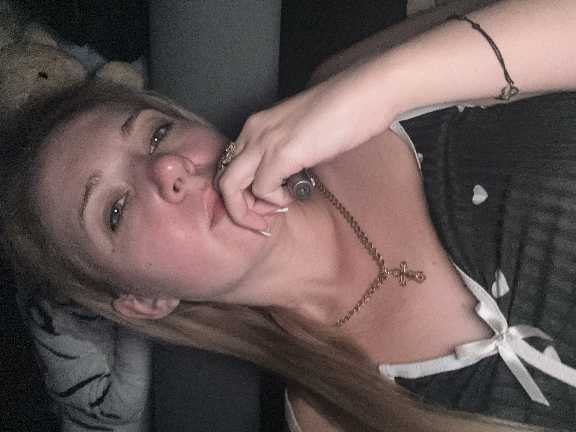 A blonde woman is lying down, with her head tilted slightly. Her right hand is raised to her mouth, with her fingers resting on her chin. She is wearing a dark, lacy top with a white bow in the center. A gold chain with a cross pendant is visible around her neck, and a dark bracelet with a small heart charm is on her right wrist. The background is dark, with hints of bedding and a stuffed animal visible. The scene appears to be indoors, in a dimly lit bedroom.
