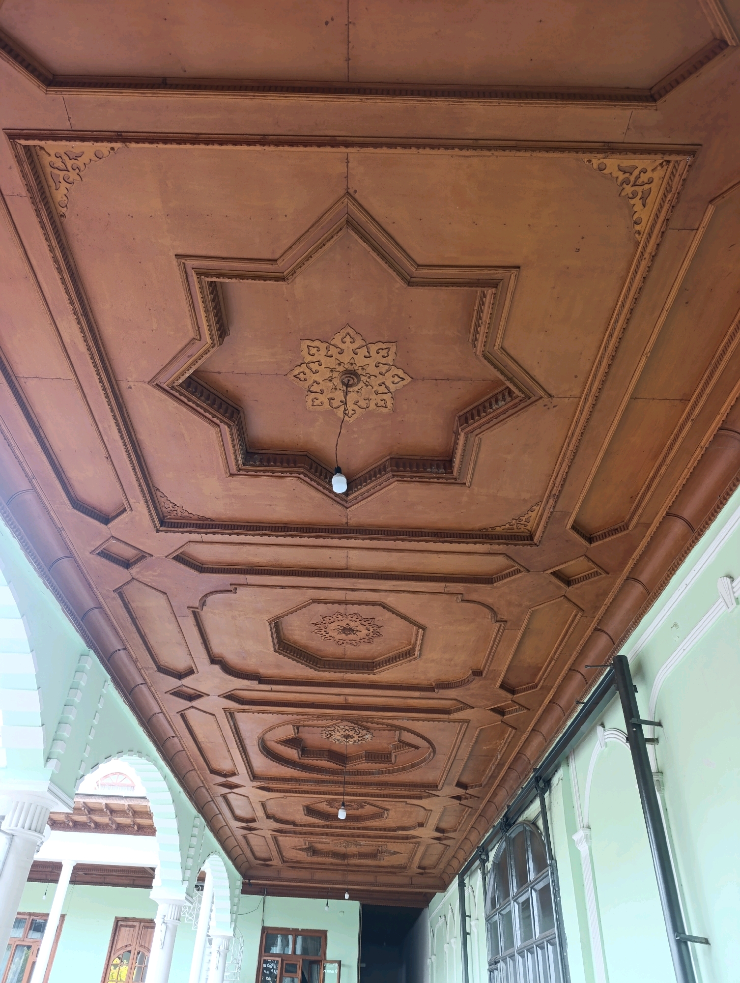 The image shows a wooden ceiling with intricate geometric carvings in Margilan City, Uzbekistan. The ceiling is coffered, with each coffer featuring carved motifs, including an eight-pointed star in the center of the frame. The wood appears to be dark brown and aged. Several light fixtures hang from the ceiling. The ceiling is viewed from below, looking upwards, and extends across a covered walkway or arcade. On the right side of the frame, a light green wall with white trim and arched window openings is visible. A dark metal pole is also present on the right, attached to the wall and extending upwards. On the left side of the frame, architectural elements of the arcade, including columns and arches, are visible, also painted in light green with white trim. The overall impression is of traditional Uzbek architecture with a focus on detailed woodwork.