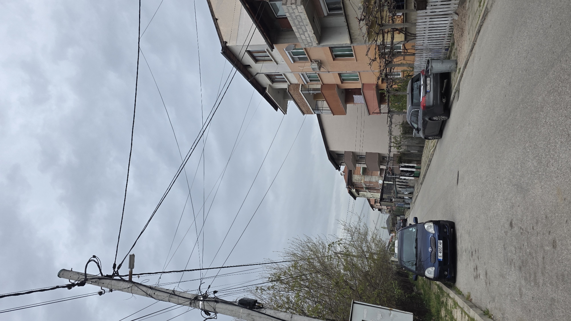 The image depicts a street scene in Blagoevgrad, Bulgaria. The viewpoint is from the side of a street, looking along its length. A blue Toyota Yaris is parked on the right side of the road, facing the camera. Further up the street, also on the right side, a dark gray Opel is parked. Buildings line the left side of the street, including a medium-rise apartment block with multiple windows and balconies. Overhead, power lines crisscross the sky. A concrete utility pole is visible on the left, supporting the power lines. The sky is overcast with grey clouds. A leafless tree is also present on the left side of the street, near the Toyota. The road is asphalt and appears to be in moderate condition. A partial view of a sign is present on the left side, in the lower part of the frame.