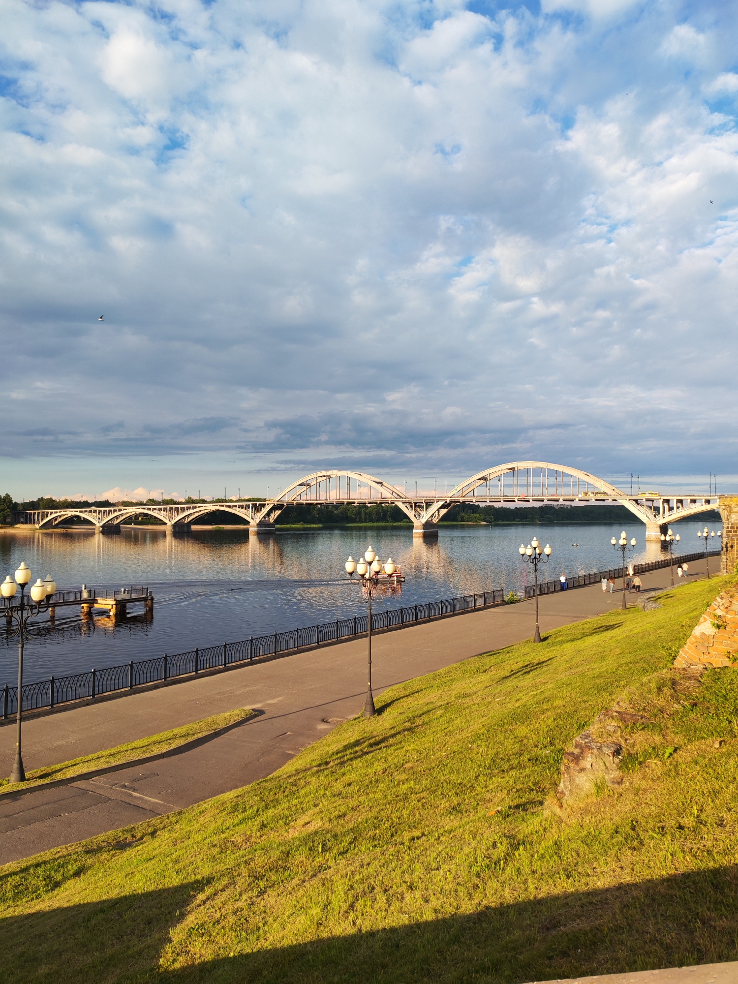 This media file depicts a scenic view of a wide river with a grand arch bridge spanning its width. The bridge, with its white arches and supporting pillars, dominates the middle ground of the image. In the foreground, a paved walkway runs alongside the river, bordered by a black, ornate metal fence. Tall, old-fashioned lampposts line the walkway, some of which are lit. To the right of the walkway, a grassy embankment slopes upwards, casting long shadows, suggesting it is either early morning or late afternoon.
The river itself is calm and reflects the bright sky and the bridge. On the water, a small pier extends from the bank, with what appear to be a few small boats or floating structures tied to it. A few distant figures are visible on the walkway, strolling and enjoying the view.
The sky is a mix of blue and scattered white and gray clouds, with a hint of sunlight breaking through, creating a pleasant, somewhat dramatic atmosphere. A single bird is visible flying high in the sky, adding a touch of life to the expansive scene. The overall impression is one of a tranquil and picturesque riverside setting, possibly a park or promenade area, in Bol'shie Mkhi, Russia. The time of day appears to be either early morning or late afternoon due to the long shadows and the quality of light. There is no visible text in the image.