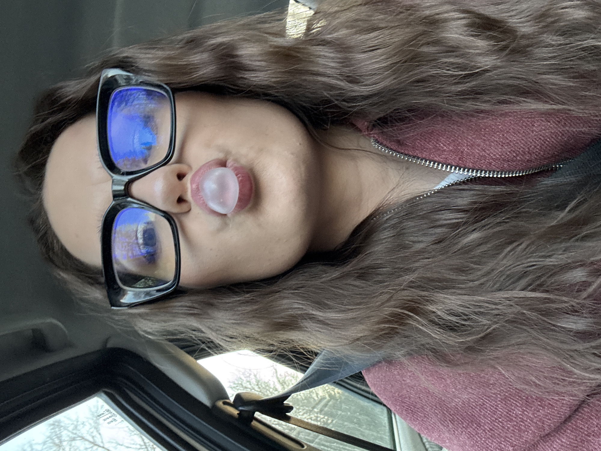 A person is in a car, blowing a bubble with chewing gum. They are wearing glasses and a burgundy jacket. The location is not possible to determine.