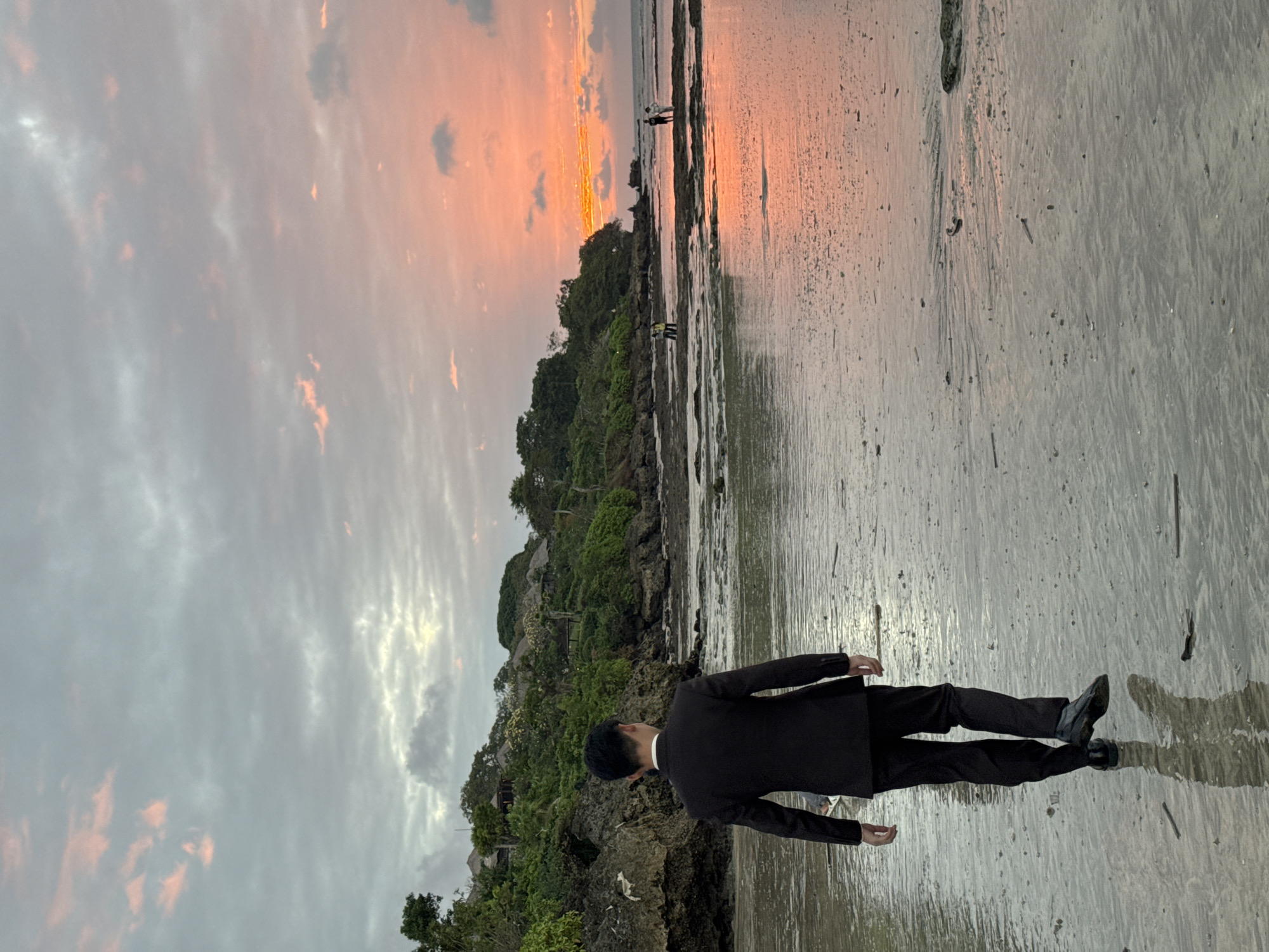 A full shot captures a man in a black suit walking away from the camera on a wet, sandy beach at dusk. The man's back is to the viewer as he walks towards a body of water. His dark hair is short and neat. He is wearing a white collared shirt underneath the suit jacket. He has on black dress shoes. The beach is littered with small pieces of driftwood and debris. The water's surface reflects the warm, orange and pink hues of the sunset sky.
In the background, lush green trees and vegetation cover a rocky hillside. Several small buildings with thatched roofs are nestled amongst the foliage. To the right of the frame, a rocky shoreline extends into the sea. A few other individuals are visible in the distance, walking along the water's edge. The sky above is a mix of gray clouds tinged with the colors of the sunset. The overall scene suggests a tranquil evening on a tropical coast, possibly in the vicinity of Kuta Selatan, Indonesia, known for its picturesque beaches and resorts.