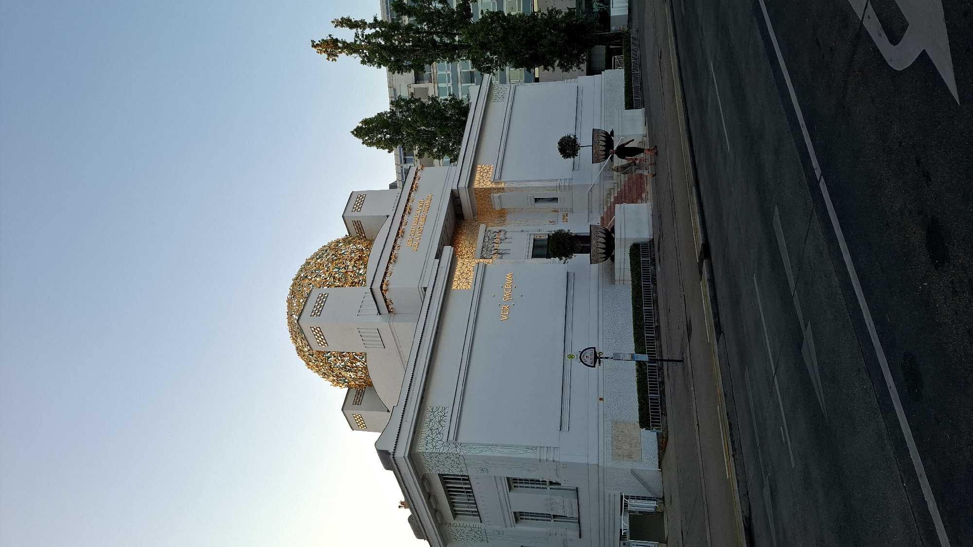 The image shows the Secession Building, an art nouveau exhibition hall located in Vienna, Austria. The building is white with a distinctive golden dome composed of laurel leaves. The facade features elaborate golden ornamentation and the inscription "Der Zeit ihre Kunst, der Kunst ihre Freiheit" (To every age its art, to every art its freedom). Two individuals are walking up the steps towards the entrance, and two trees in planters flank the entrance. A street with traffic markings is in the foreground. Behind the Secession Building, modern high-rise buildings are visible, along with tall trees. The sky is clear and blue, suggesting daylight.