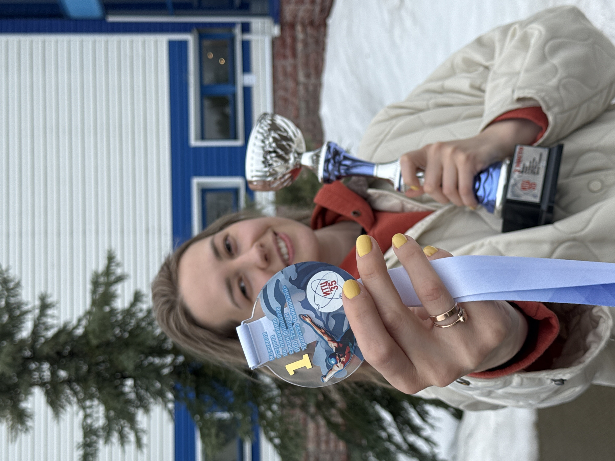 A young woman is standing outside in a snowy environment, holding a trophy and a medal. The medal is clear with a blue design, featuring the number "1" in gold, and the text "Матчевая встреча по плаванию Среди филиалов 000 'Газпром Трансгаз Чайковский' ИТЦ 35," suggesting a swimming competition among branches of Gazprom Transgaz in Chaikovsky. The woman is wearing a cream-colored quilted jacket with a red hoodie underneath, has yellow nail polish, and is smiling. The background shows a white and blue building, likely in the city of Chaikovsky, Russia, given the Russian text on the medal.