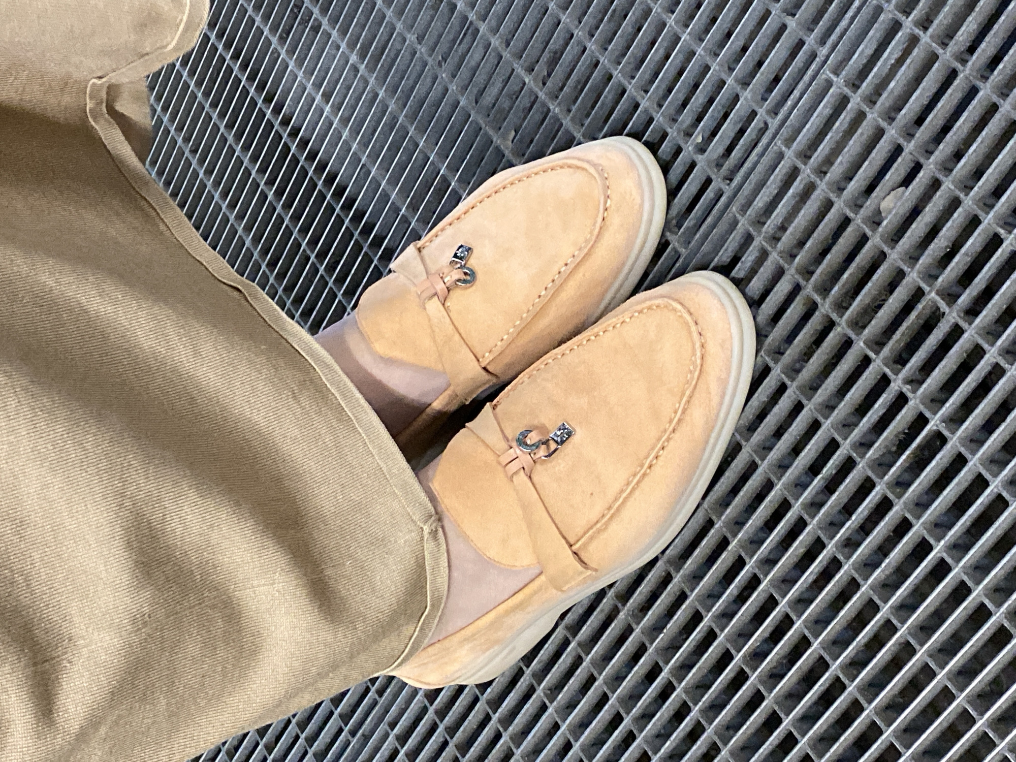 A pair of peach-colored suede loafers with metal charms are worn by a person, whose beige pants cover their ankles, and are placed on a metal grate sidewalk. The image does not provide enough information to determine a specific location in Yekaterinburg, Russia.