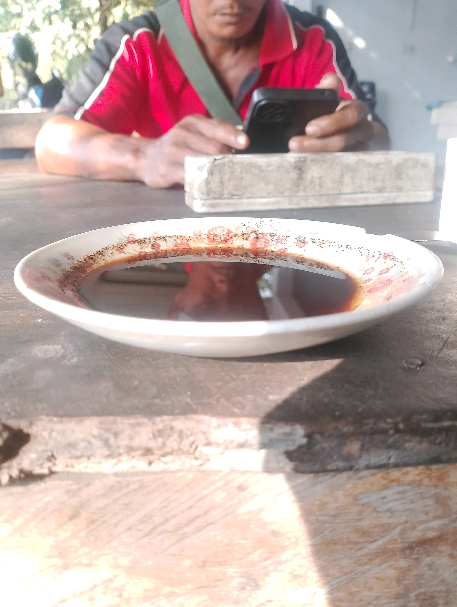In Jambean, Indonesia, a close-up shot focuses on a bowl of dark liquid with small dark particles on its rim, resting on a weathered wooden surface. In the background, a person wearing a red and black shirt and a green strap across their chest is blurred, holding a black smartphone. The scene appears to be outdoors, with natural light casting shadows.