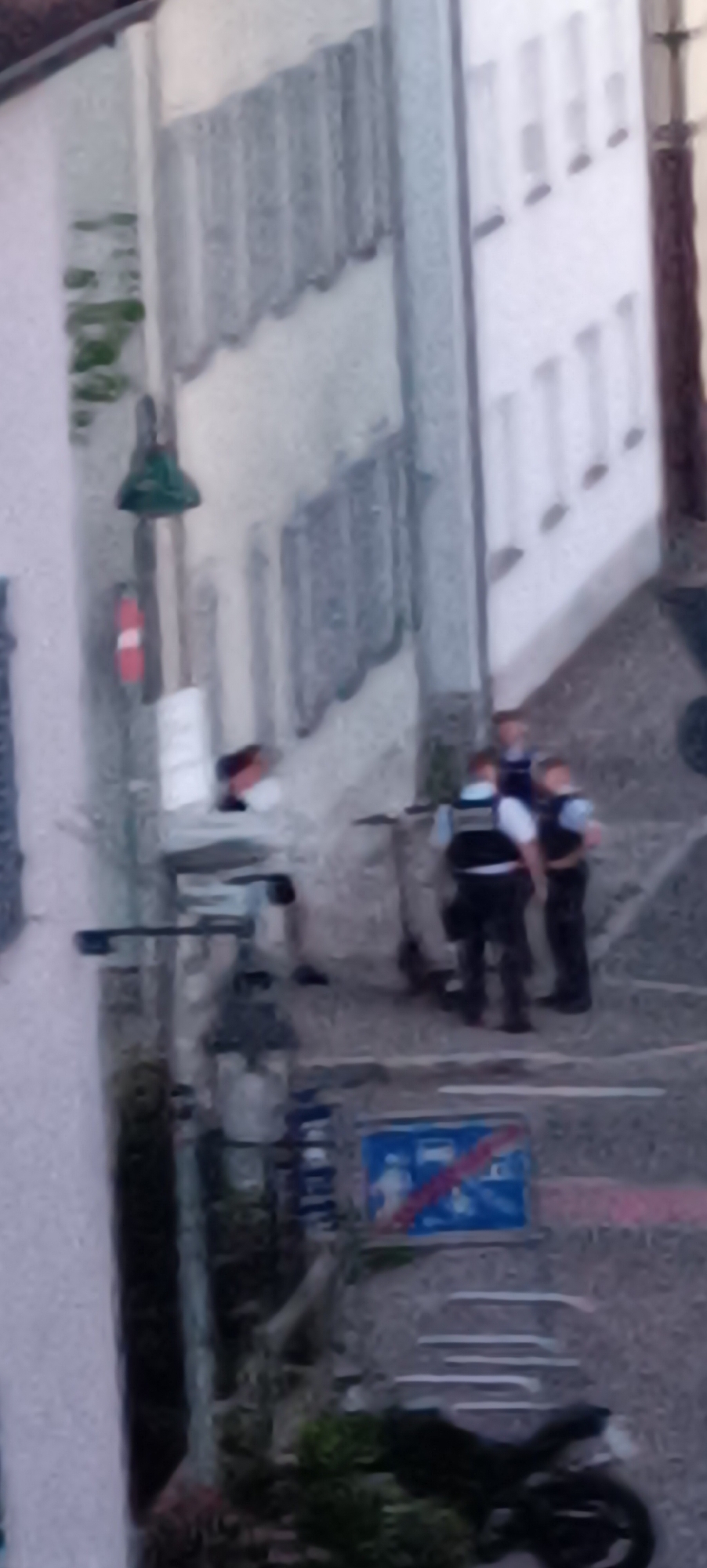 The image depicts a street scene in Winnenden, Germany. In the foreground, there is a motorcycle parked on the right side of the image. A blue sign with red diagonal lines across it is visible in the lower middle of the frame, indicating a prohibition. Above this, a group of people are gathered, some of whom appear to be wearing dark uniforms with vests, possibly law enforcement officers. Two individuals, one in the foreground and another slightly behind, are standing near a scooter or electric bike. Further in the background, a building with multiple windows is visible, and to the left, another building with an awning. Green foliage is also present in the scene.