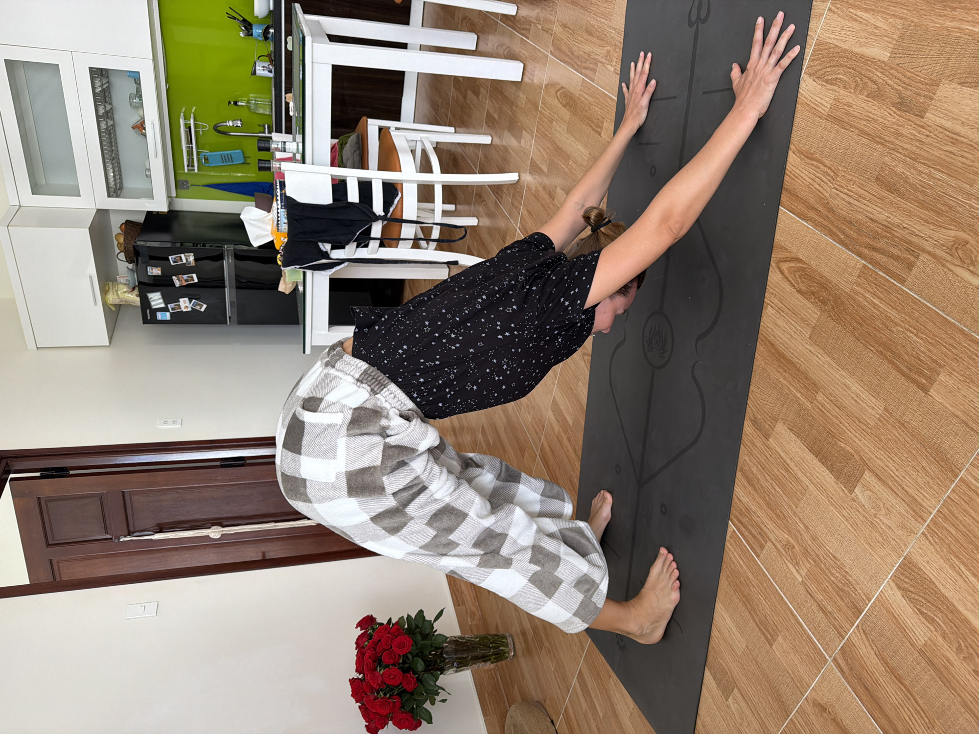 The image depicts a woman performing a downward-facing dog yoga pose on a dark yoga mat placed on a wooden-tiled floor. She is wearing loose-fitting checkered pants and a black top with white star patterns. To the left of the woman, a vase filled with red roses is visible. In the background, a partially open wooden door, a refrigerator, kitchen cabinets, and a white dining table with chairs are present. The scene appears to be an indoor residential setting, possibly a kitchen or dining area, located in Lâm Viên - Đà Lạt, Vietnam, indicated by the presence of red roses, which are culturally significant in the region.