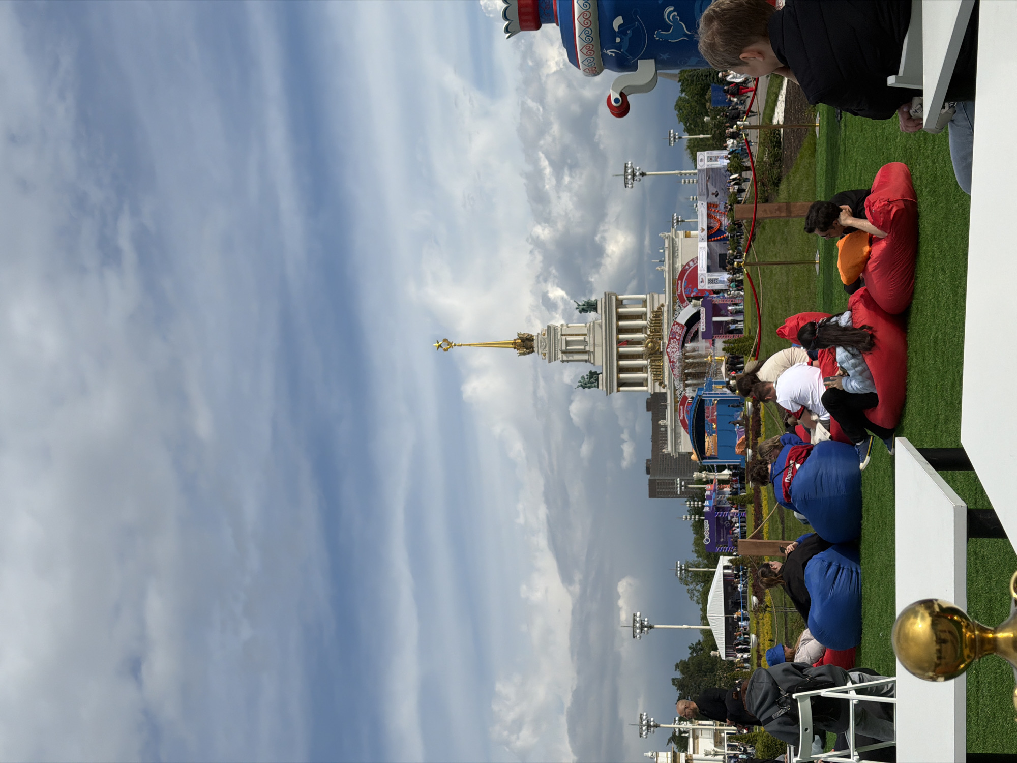 The image depicts an outdoor scene in Moscow, Russia, likely at the VDNKh (Exhibition of Achievements of National Economy) park. The iconic Central Pavilion with its golden spire is visible in the background. In the foreground, people are seated on colorful beanbag chairs scattered on a green lawn. They appear to be watching something, possibly a screen or a performance, though the focus of their attention is not clearly visible. Some people are sitting and resting while others are looking at their phones. The sky is cloudy, with shades of blue and grey. Street lamps and decorations are scattered throughout the area. Part of a large, ornate blue and white decorative object is visible on the upper right.