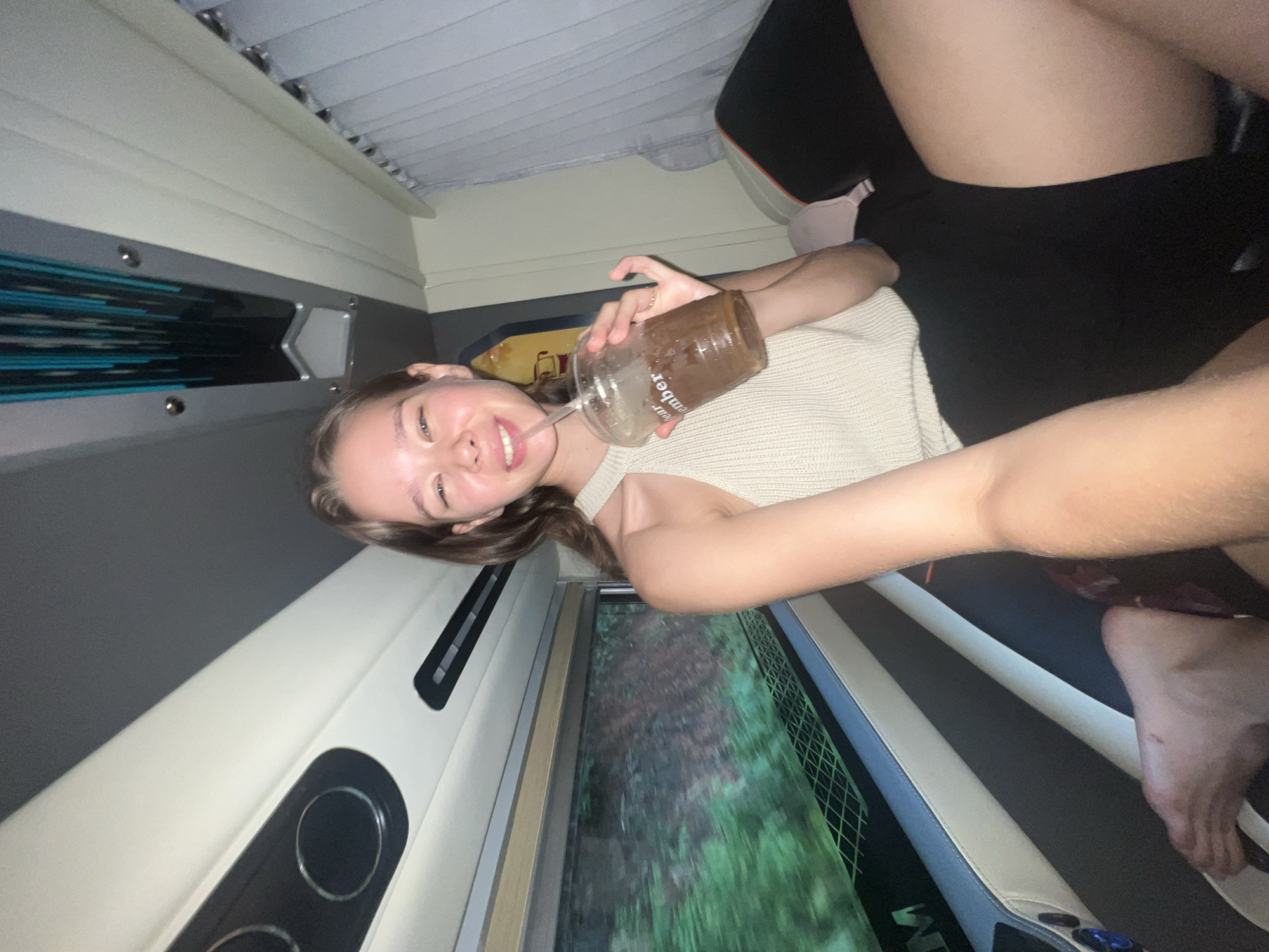 A young woman is smiling brightly, captured in a rotated close-up perspective while relaxing inside what appears to be a sleeper bus or van, suggested to be in Bo Y, Vietnam. She is lying down, wearing a light-colored, possibly knitted, tank top and dark shorts.
She holds a clear plastic cup with a straw, containing a dark beverage that resembles coffee or a chocolate drink. The word "December" is visible on the side of the cup. Her expression is cheerful and relaxed.
The interior of the vehicle features light-colored paneled walls and ceiling, with some darker grey and blue accent elements along the side. Through a window behind her, a blurred view of lush green foliage is visible, indicating that the vehicle is in motion and traveling through a natural, outdoor landscape. A pink bra is partially visible near her leg. The lower part of the interior, possibly a seat or bed, has stripes of light and dark material. The time of day is not explicitly clear but the blurred greenery suggests daytime travel.