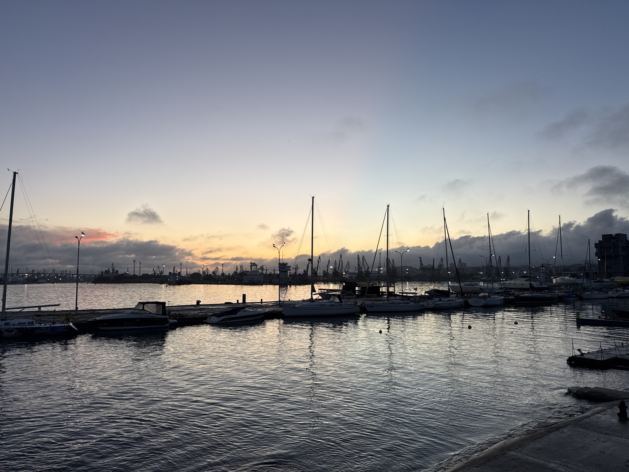 The image captures a serene view of a marina or harbor at either sunrise or sunset, characterized by the warm, gradient sky. The foreground is dominated by calm, dark water with gentle ripples reflecting the soft light. Several boats, primarily sailboats with tall masts and some motorboats, are docked along wooden piers on the left and center.
In the mid-ground, the water surface extends towards a distant shoreline. The background reveals an active industrial port with numerous large cranes, some illuminated by artificial light, and various structures suggesting commercial shipping operations. A bridge or elevated road is also faintly visible in the distance.
The sky transitions from a soft blue-grey at the top, through purples and pinks, to a vibrant orange and yellow glow near the horizon, indicative of the golden hour. Patches of dark clouds are scattered across the sky, particularly on the right and above the industrial area, contrasting with the brighter colors. No people are visible in the scene.
Notable details include lampposts on the docks, some of which are lit. Faint text "Lady Blue" is visible on the bow of a smaller boat on the far left, and "Merlin" is discernible on a white motorboat docked towards the center-left. The overall atmosphere is peaceful, blending natural beauty with industrial activity under a dramatic sky.
