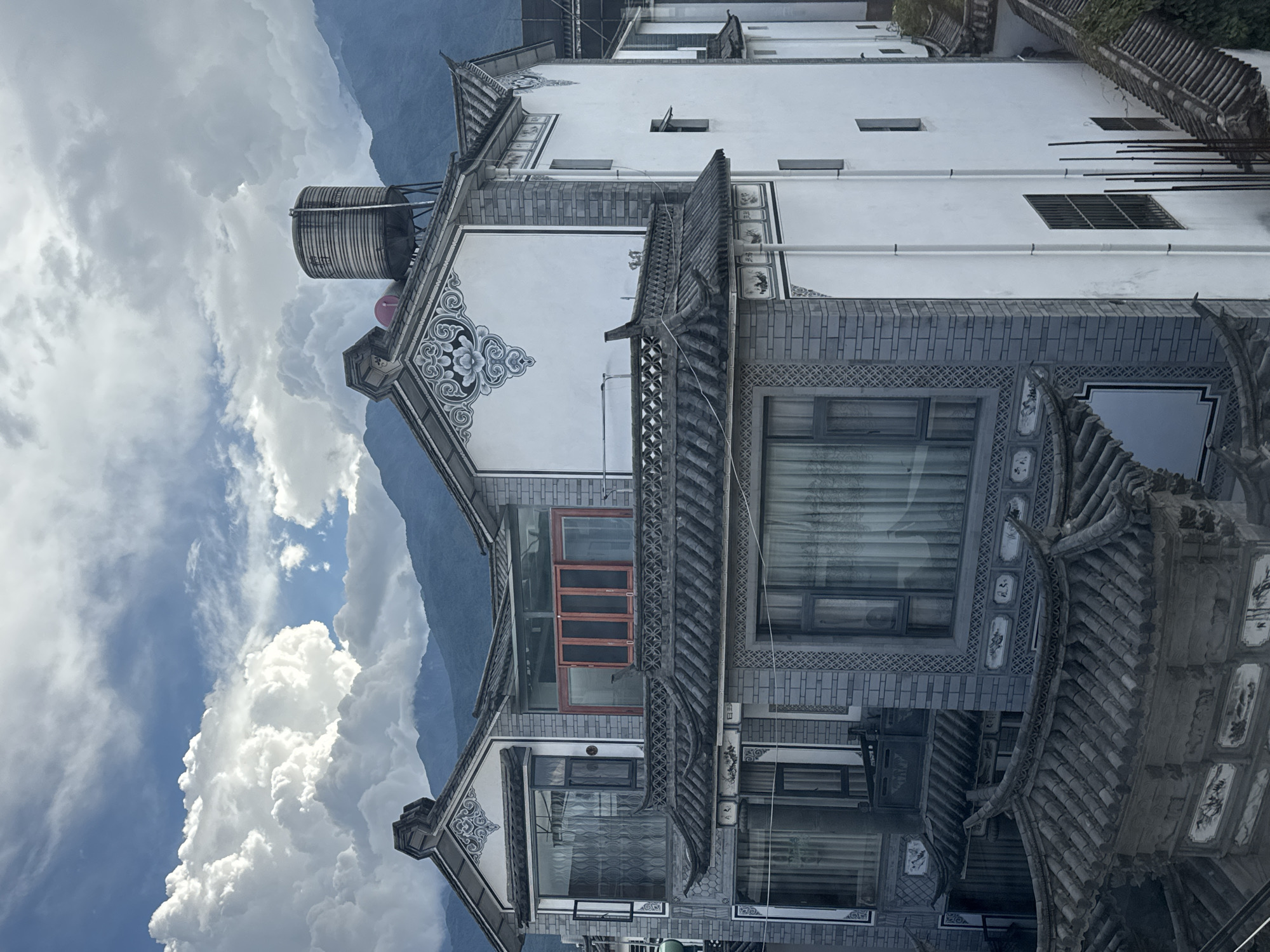 The image shows a multi-story building with traditional Chinese architecture, located in Dali City, China. The building features grey tiled roofs with curved eaves, white walls with decorative elements, and grey brickwork. Windows are visible on all levels, some with sheer curtains. On the rooftop, a large cylindrical water tank is present. In the background, rolling mountains are shrouded in clouds. The sky is partly cloudy with dramatic, fluffy white clouds. The scene is captured from an elevated viewpoint, looking down and across the building.