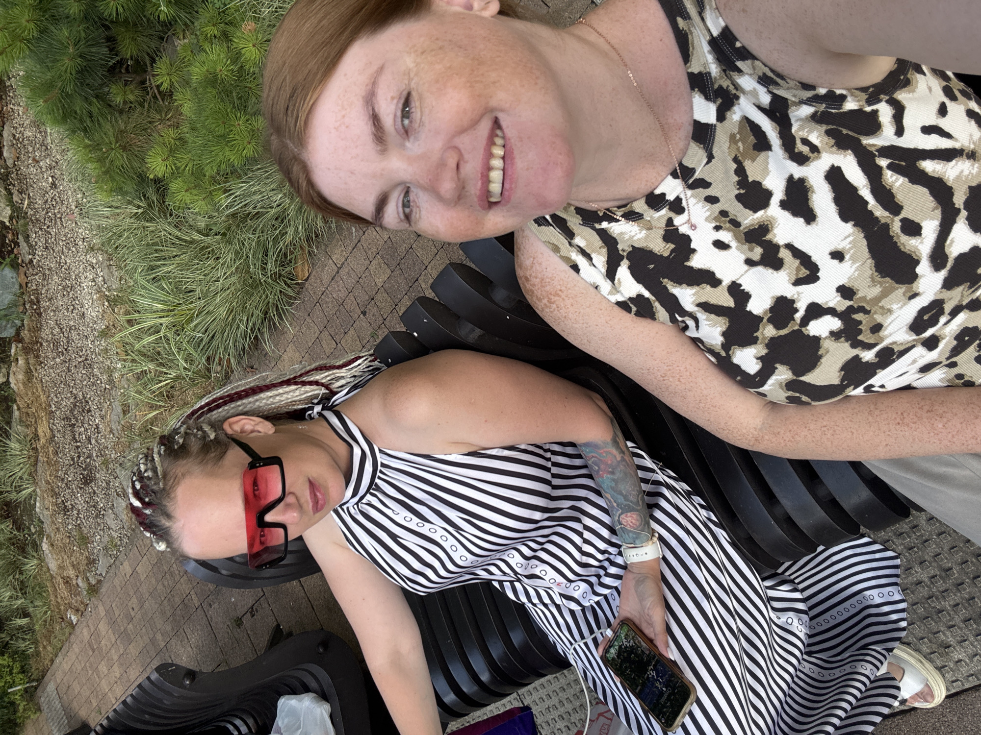 Two women are sitting on a black slatted bench in an outdoor setting. The woman on the left has long, braided hair with blonde, red, and dark brown streaks. She is wearing oversized pink square sunglasses and a black and white striped halter dress. She has a colorful tattoo on her left arm and is holding a smartphone in her right hand. The woman on the right has short, reddish-brown hair and is smiling. She is wearing a sleeveless top with a black, white, and tan leopard print pattern. Both women are looking towards the camera, suggesting they are taking a selfie. The background shows greenery, gravel, and brick paving, indicating an outdoor park or garden. The lighting suggests it is daytime, likely sunny. There is no visible text in the image. The context provided indicates the location is Sochi, Russia.