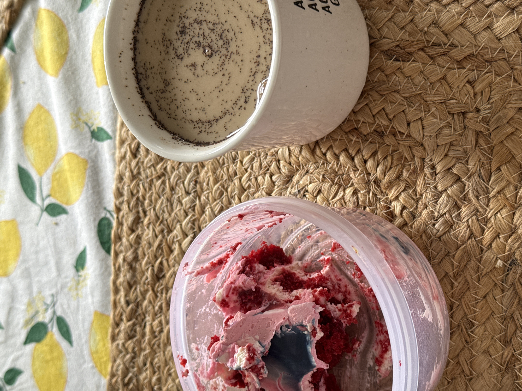 On a woven placemat sits a mug of coffee with poppy seeds and a jar of leftover red velvet cake with pink frosting. A lemon-patterned napkin is visible on the left. Location is uncertain.