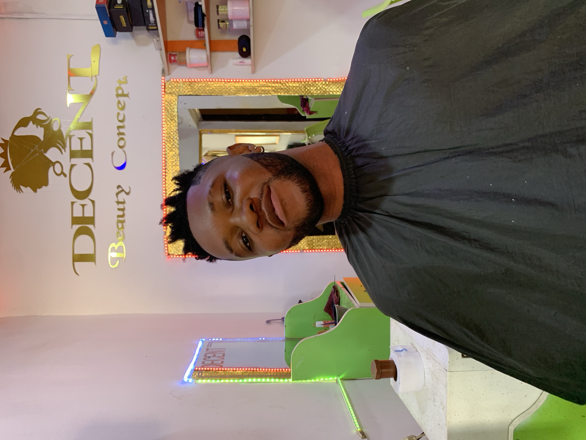 A man with a freshly trimmed haircut and beard is wearing a black cape. He is in a hair salon called "Decent Beauty Concept" located in Ikorodu, Nigeria. The salon features mirrors with LED lighting, gold lettering on the wall, and shelving with various hair products. The background includes an orange shelf with miscellaneous items.