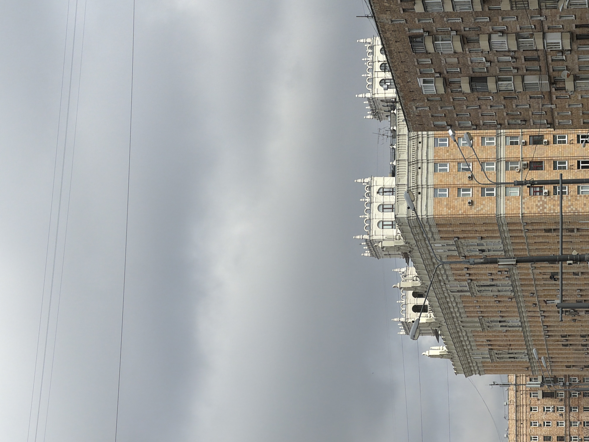 Here is a description of the image:
The image shows a tall building in Moscow, Russia under a cloudy sky. The building appears to be made of brick with white architectural details on the upper floors. Power lines are visible in the foreground against the sky. The photograph seems to have been taken at an angle, looking upwards towards the building. The architecture and the cloudy sky suggest a typical urban scene in Moscow.