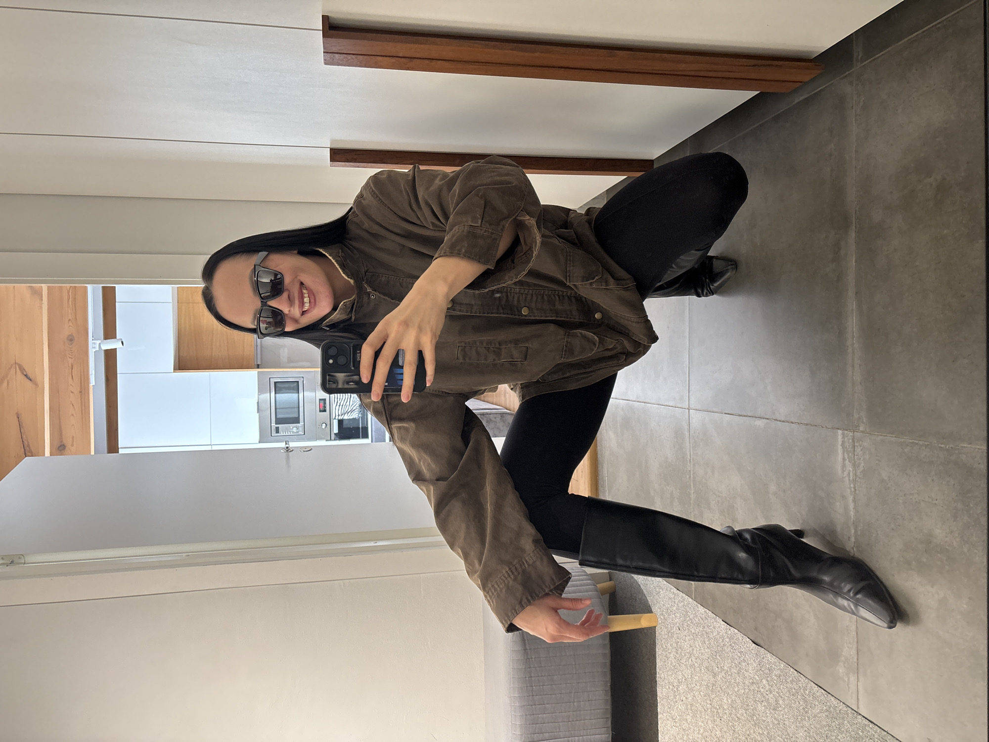 The image displays an indoor scene identified as located in the city of Chaikovsky, Russia, within what appears to be a modern residential setting. A female individual, with long dark hair, is captured in a squatting position while taking a selfie with a black smartphone held in her right hand. She is wearing black sunglasses, a brown, collared utility-style jacket with multiple buttons and pockets, black leggings, and black, high-heeled, pointed-toe boots. Her left hand rests on her left thigh, and she is smiling broadly, revealing her teeth.
The flooring in the foreground is composed of large grey tiles, transitioning to a light grey carpeted area on the left. On this carpet, a light grey upholstered bench with light wooden legs is partially visible. The walls are predominantly white, with vertical dark wooden trim panels positioned on the right side of the frame. In the background, beyond a doorway, an open-plan kitchen area is discernible, featuring white upper and lower cabinetry, a built-in stainless steel microwave oven, and an oven unit situated below it. The ceiling in this background area showcases exposed wooden beams and a white linear light fixture. The environment is brightly and evenly lit.