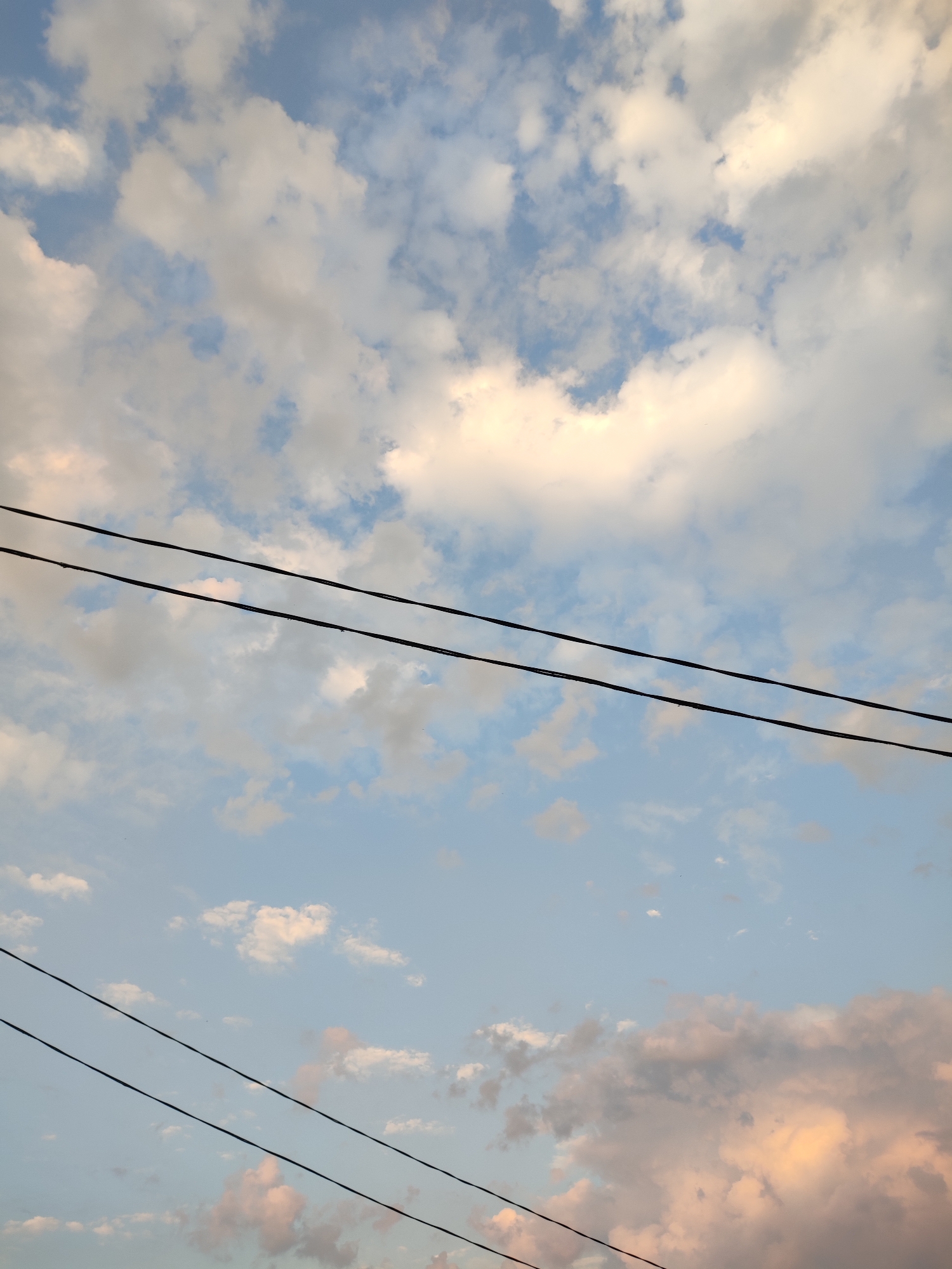 This is a vertical, medium-angle outdoor photograph of a pale blue sky filled with scattered white and grey clouds. Three black utility wires are diagonally stretched across the frame from the upper left to the lower right. The wires are parallel to each other. Some clouds are tinged with a pale yellow and pink, suggesting it might be dawn or dusk. The scene is devoid of any people or structures, indicating it is a view of the sky. This image was taken in or near Варнянскі сельскі Савет, Belarus.