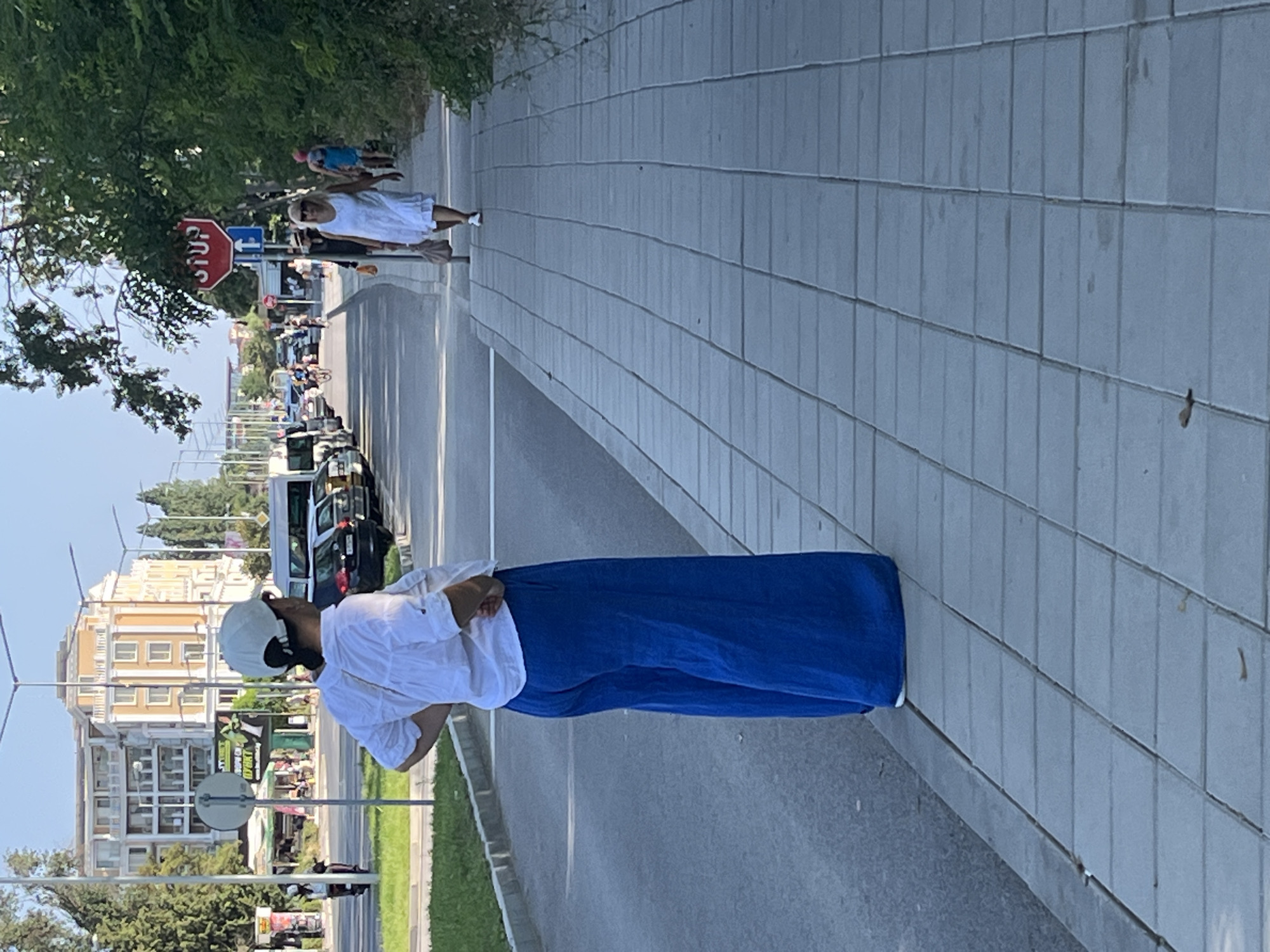 A woman in a white shirt and wide blue pants stands on a sidewalk in Nesebar, Bulgaria. She is facing away from the camera, with her hands on her hips. In the background, a street is visible with cars and other pedestrians. A multi-story building with yellow accents is on the left side of the frame, and lush green trees line the right side. A red octagonal stop sign is posted on a pole on the right side of the street. The sidewalk is paved with rectangular concrete tiles.