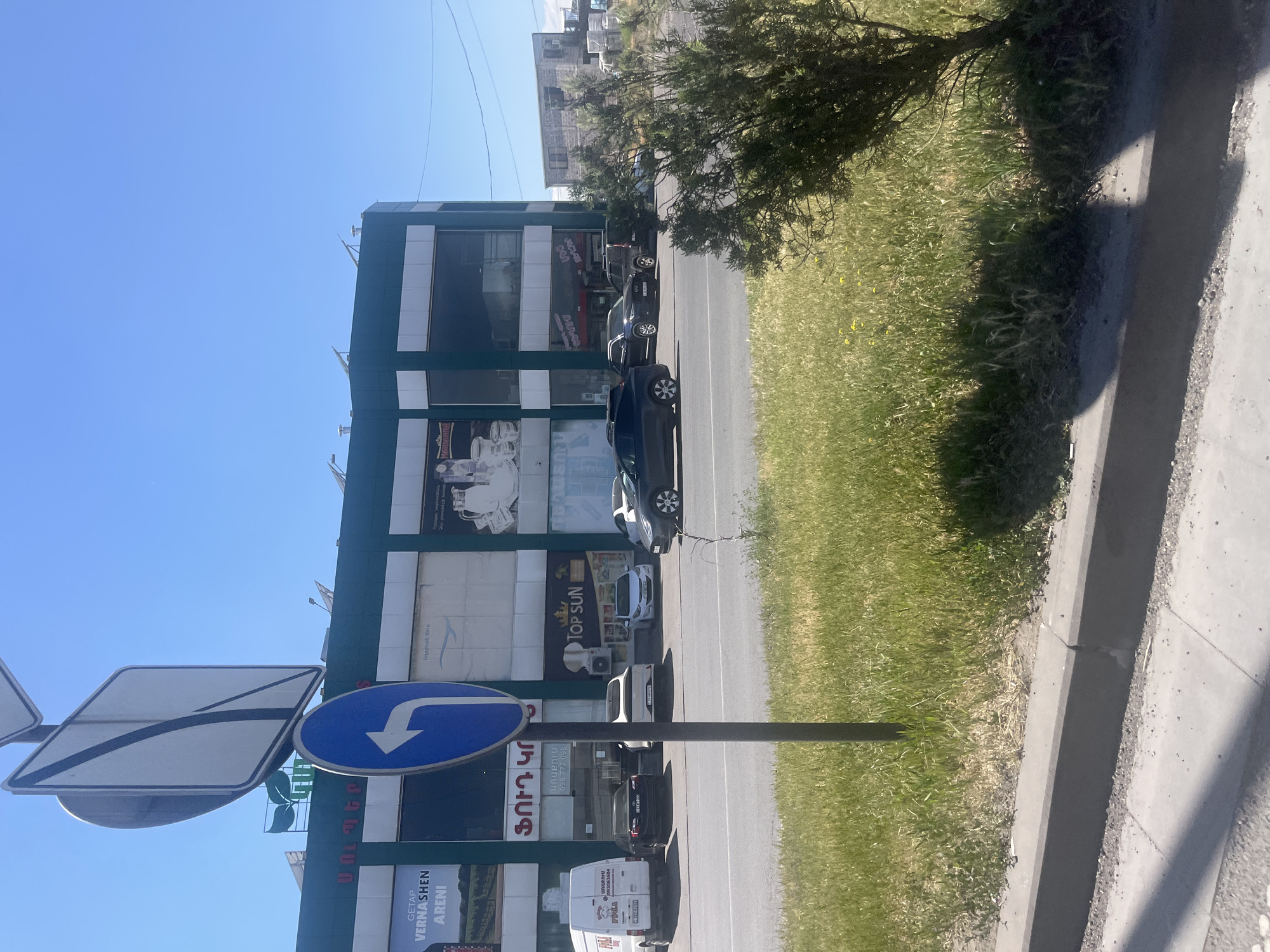 The image captures an urban street scene in Yerevan, Armenia, on a clear, sunny day. The dominant feature is a multi-story commercial building with a dark facade and large windows, occupying the left side of the frame. Several cars are parked along the street in front of this building, including a white van, a black SUV, a black sedan, a grey car, and a white car.
In the foreground, a pole supports two traffic signs: a blue circular sign indicating a mandatory left turn, and a larger square sign behind it with a dark diagonal stripe. The building's exterior features various advertisements and business names, primarily in Armenian, alongside English branding. Visible text includes "GETAP," "VERNASHEN," "ARENI," "TOP SUN," "Աղբյուրի Ջուր" (Spring Water), "ՖՈՒԴ ԿՈ" (Food Co), "ԱՌԱՔՈՒՄ" (Delivery), "ՏԵՂՈՒՄ" (On-site/Here), and "ԶԵՂՉԵՐ" (Discounts), along with phone numbers "093063604" and "098 777 098."
To the right of the parked vehicles, a strip of vibrant green grass with small yellow flowers runs alongside the asphalt road, separated by a concrete curb. Further right, the ground transitions to a concrete sidewalk. A tree with dense green foliage partially obscures the view of another building or structure in the background. The bright blue sky, intense sunlight, and sharp shadows suggest it is daytime, likely between late morning and late afternoon. There are no people visible, portraying a quiet moment on a commercial street.
