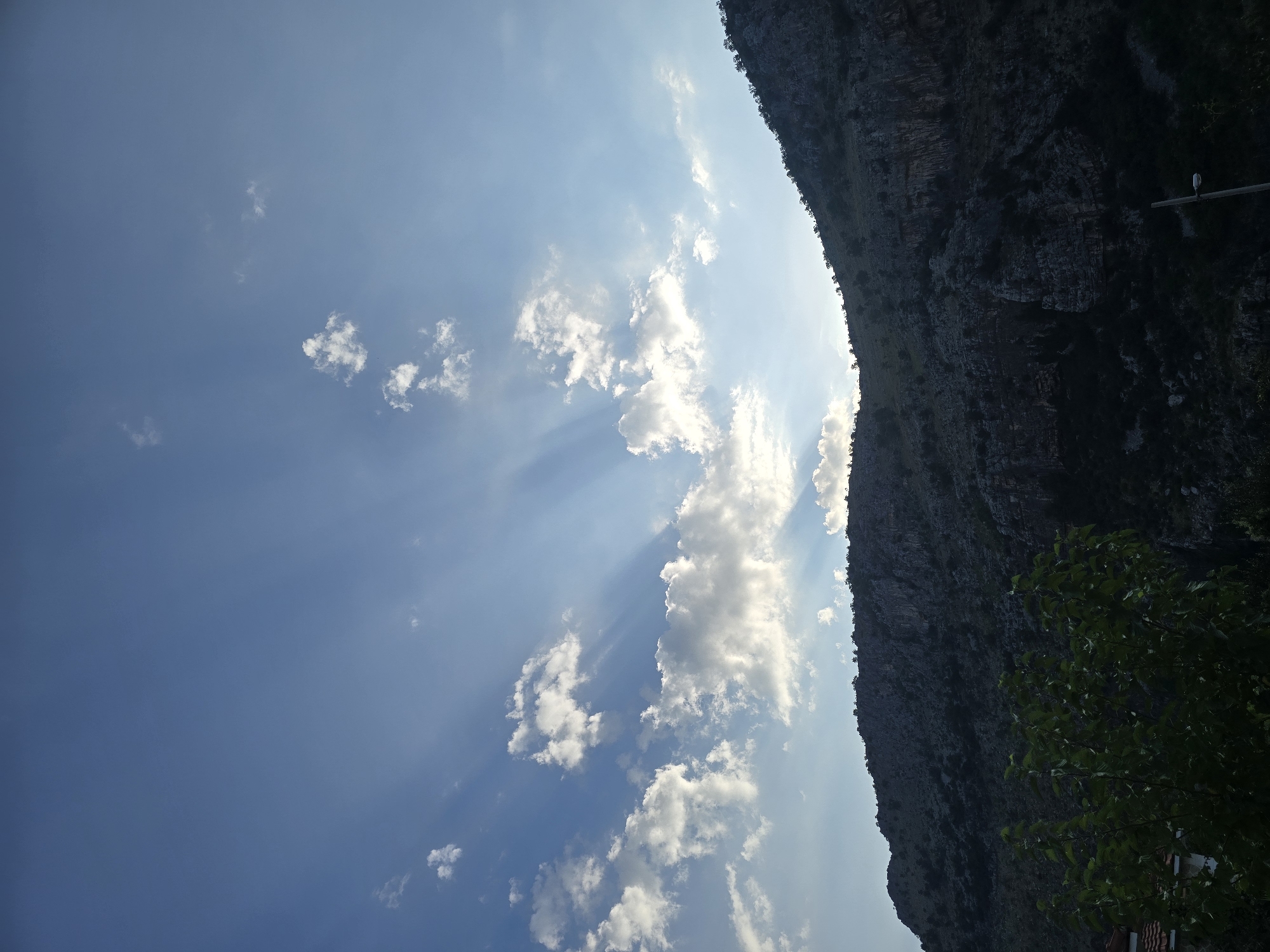 The image captures a view of a mountainous landscape with a dramatic sky. The sun is partially obscured by clouds, casting visible sunbeams through the blue sky. Scattered white clouds are present, some appearing fluffy and others more wispy. The foreground is dominated by a dark, rugged mountain slope covered in sparse vegetation and trees. The slope is textured with rock formations and shadowed areas, suggesting a rough terrain. In the lower left corner, a glimpse of green foliage from trees is visible, along with a hint of a tiled roof and a white structure. The lower right corner shows a dark silhouette, possibly more vegetation, and a tall, thin, dark pole with a light fixture near its top. The scene appears to be outdoors, likely in a natural or semi-rural setting, given the prominent mountain and vegetation. The lighting suggests it might be late afternoon or early morning, with the sun low in the sky. The location is Podgorica, Montenegro.
