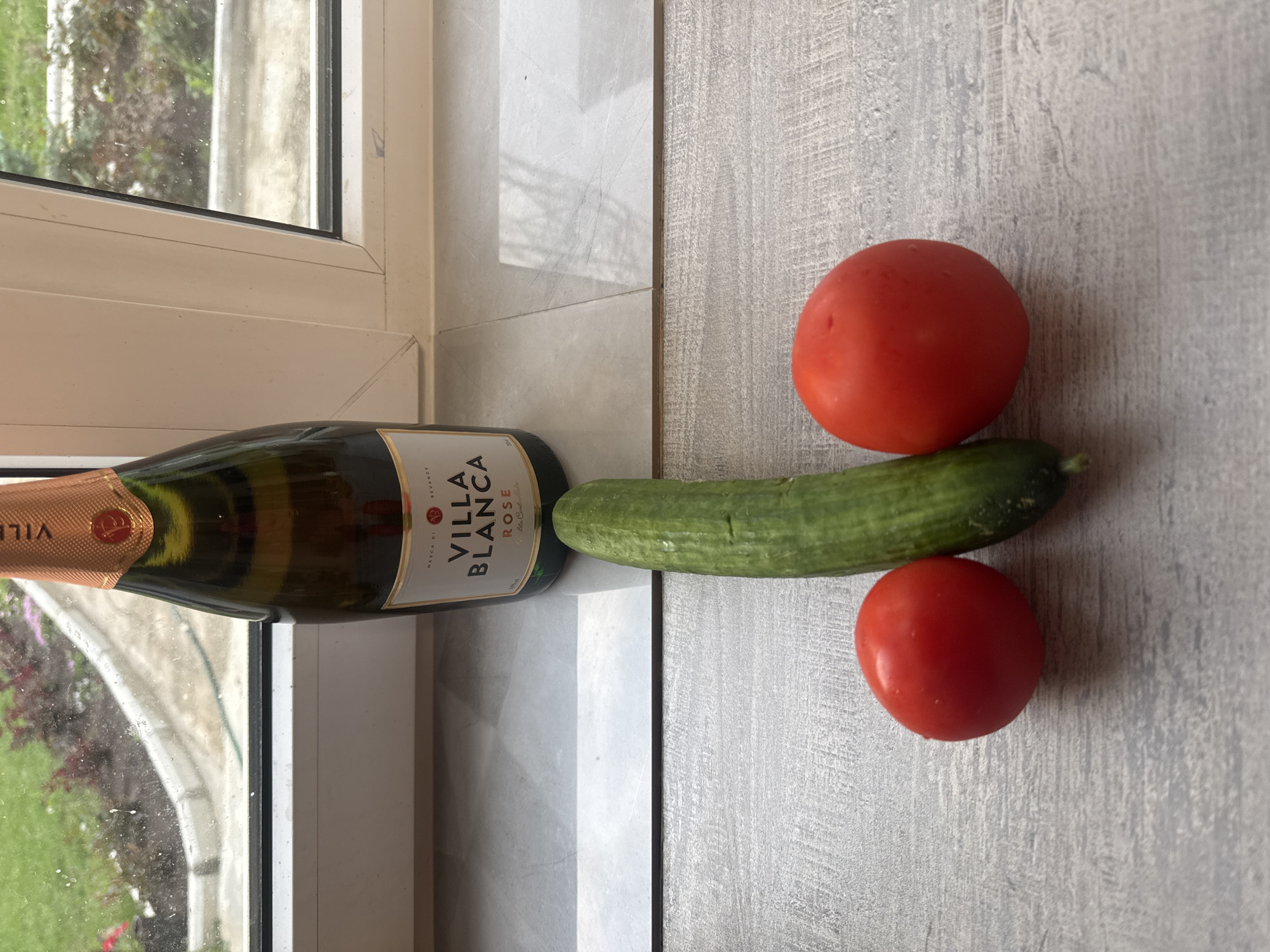 The image shows a bottle of Villa Blanca Rose sparkling wine, a cucumber, and two tomatoes lying on a surface with a wood grain texture, possibly a countertop or table. The wine bottle is near a window with a view of greenery outside. The scene is suggestive, as the arrangement of the vegetables might be interpreted as vulgar. The exact location within Chaikovsky, Russia is not discernible from the image alone.