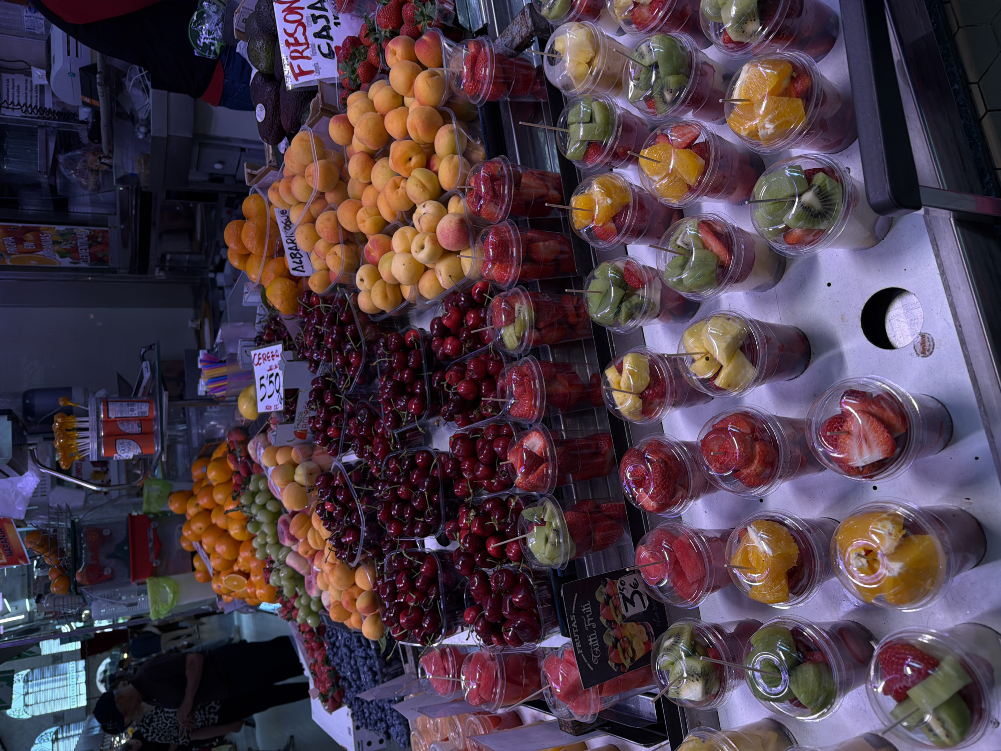 This image shows a vibrant fruit stand in the city of València, Spain. The stand is stocked with a variety of fresh fruits including apricots, cherries, oranges, grapes, strawberries, blueberries, and avocados. Some of the fruit is packaged in clear plastic cups with lids.
There are also labels indicating prices and names of the fruit: "Cereza 5,50" (Cherries 5,50), "Naranja Clementinas" (Orange Clementines), "Albaricoque" (Apricot), "Freson Caja" (Strawberry Box), "FRUTAS Tutti Frutti 3€".
In the background, a person wearing a mask can be partially seen.